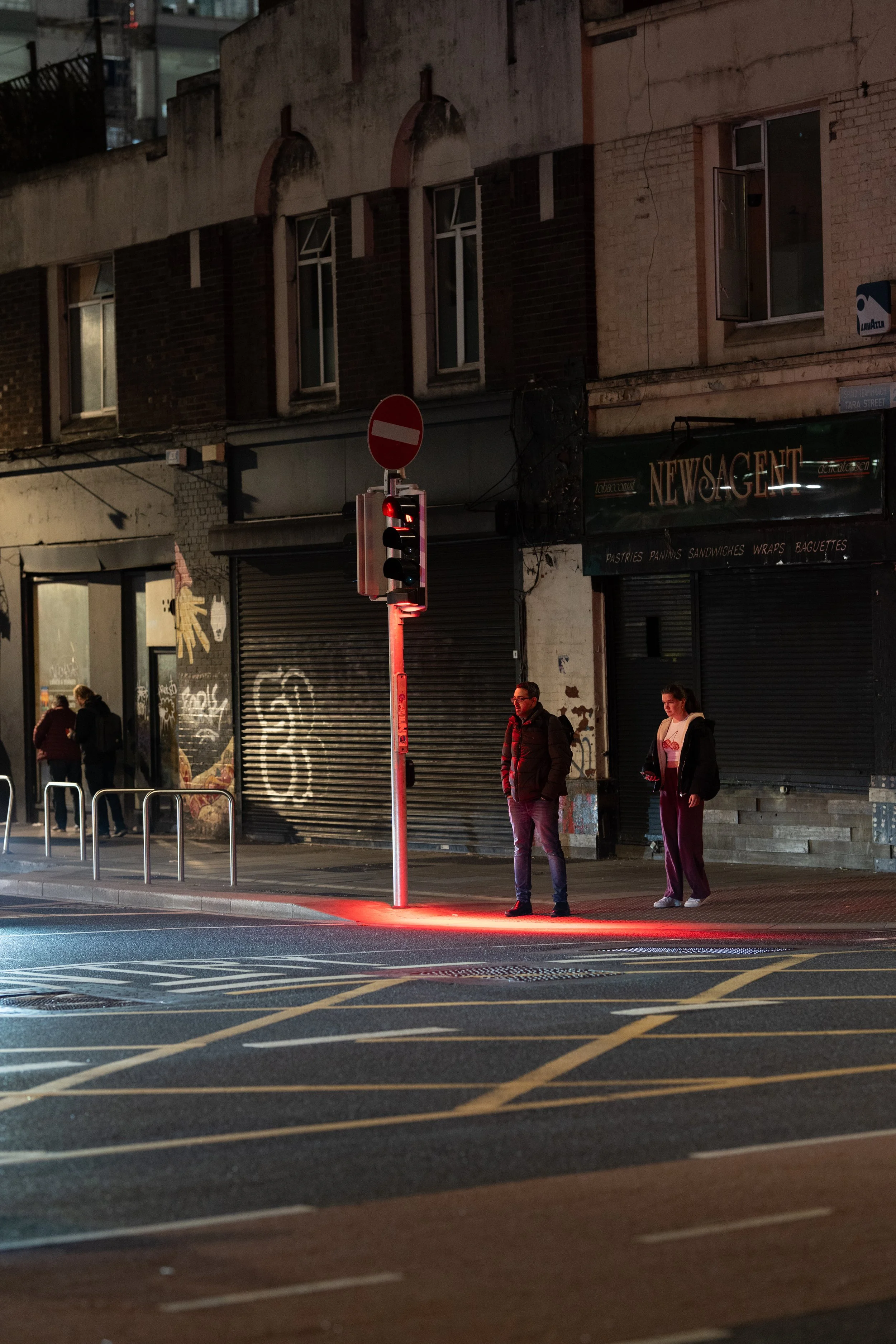 Two people standing at a crosswalk with a red traffic light in an urban area at night. The background includes a graffiti-covered wall, a closed shop with a sign 'NEWS GENT', and a dimly lit building.