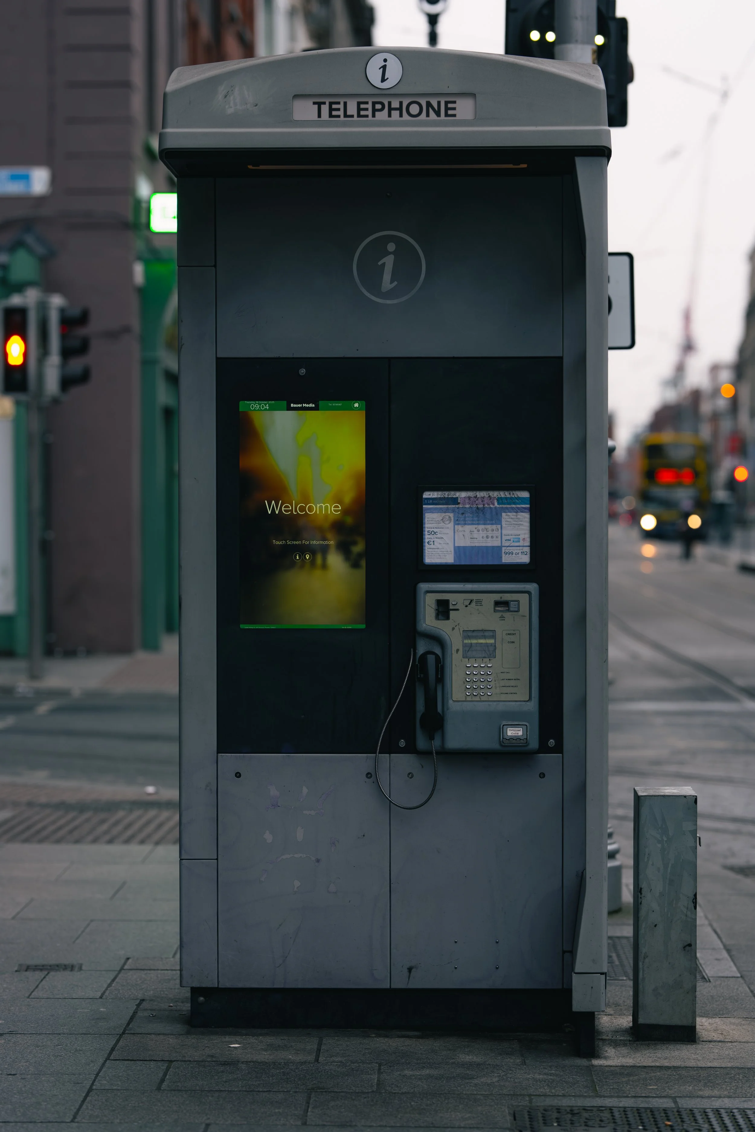 An outdoor public telephone booth with a touchscreen display showing a welcome message, a keypad, and a coin slot, situated on a city sidewalk with traffic and buildings in the background.