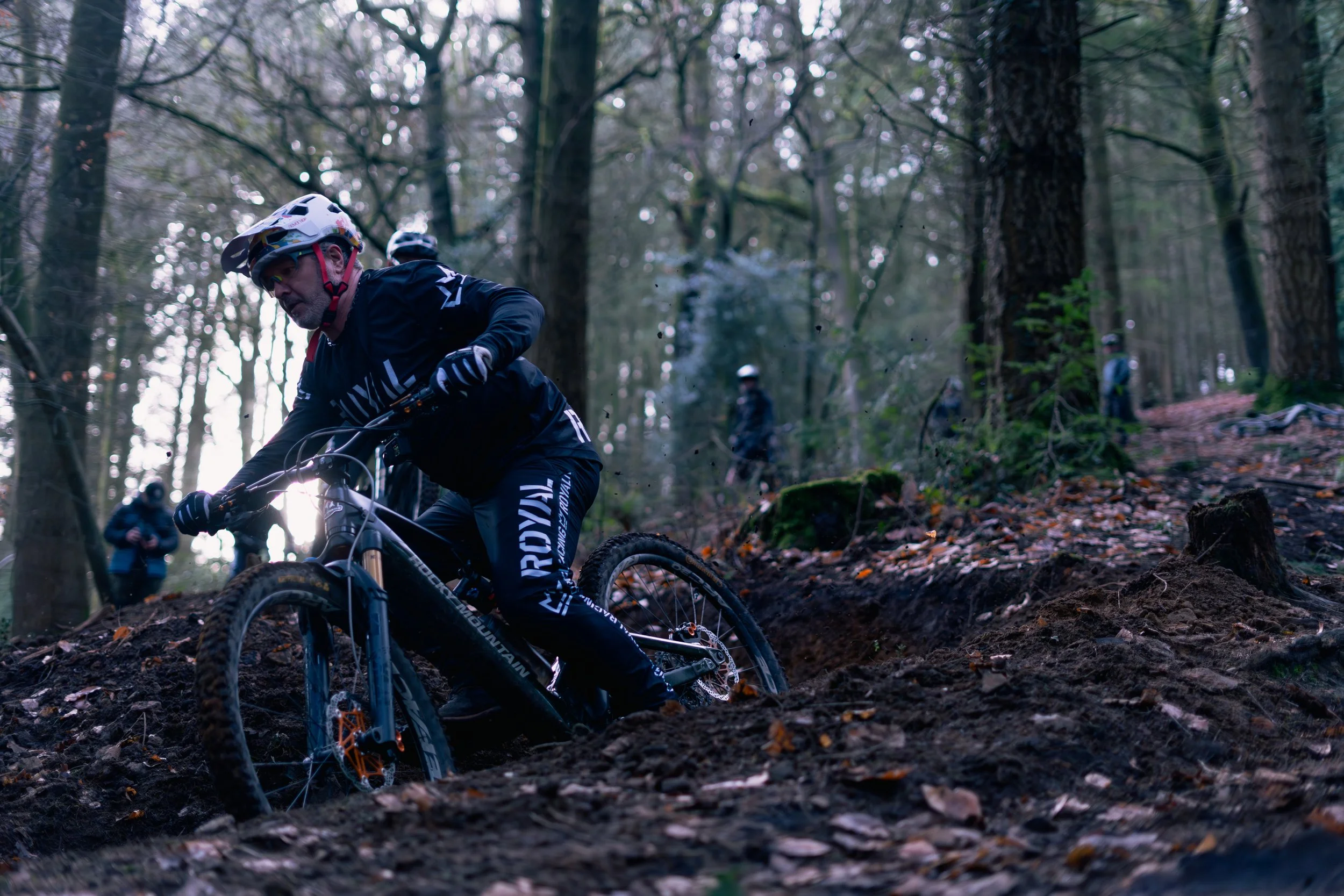 A person mountain biking on a rugged forest trail, wearing a helmet and dark sports gear, with other bikers in the background among tall trees and fallen leaves.
