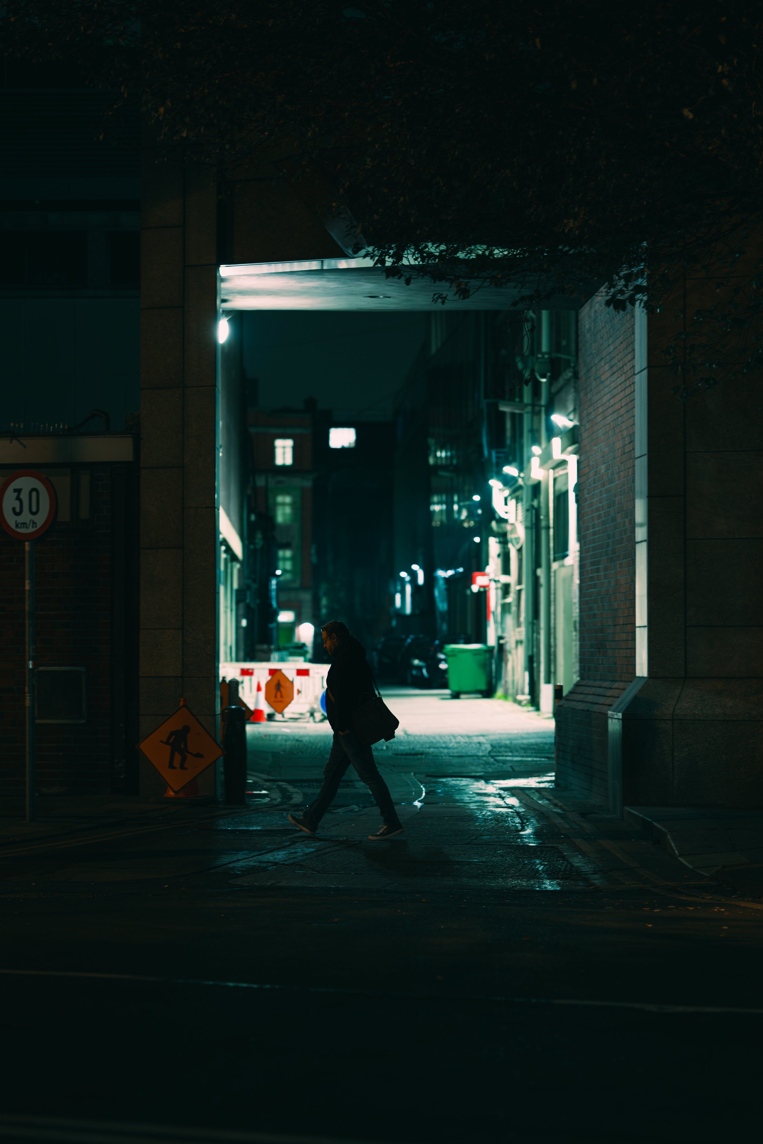 A person walking through a dimly lit city street at night with construction signs and illuminated buildings in the background.