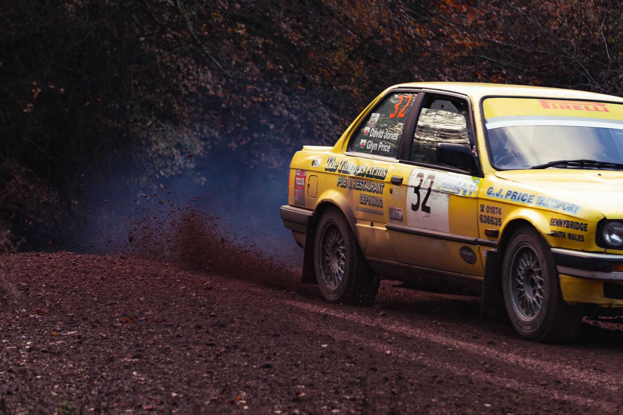 A yellow rally car with the number 32 speeds on a dirt road, kicking up dust and dirt behind it, with dark trees in the background. wye dean rally 