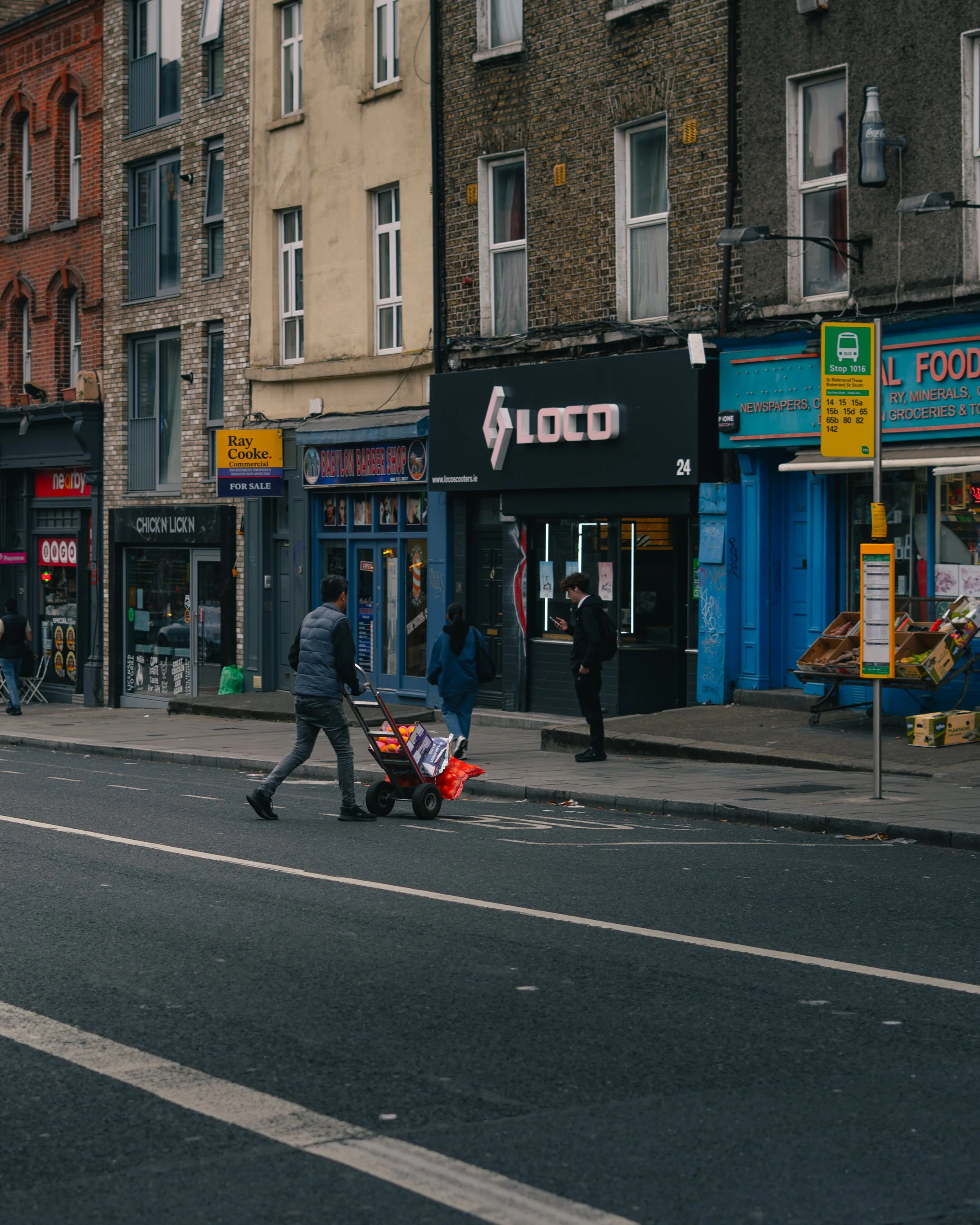 Street scene in an urban area with pedestrians, including a man pushing a shopping cart filled with oranges, crossing the street. Buildings with storefronts are visible, including a barbershop, a tech repair store, and a food market, with signs and a