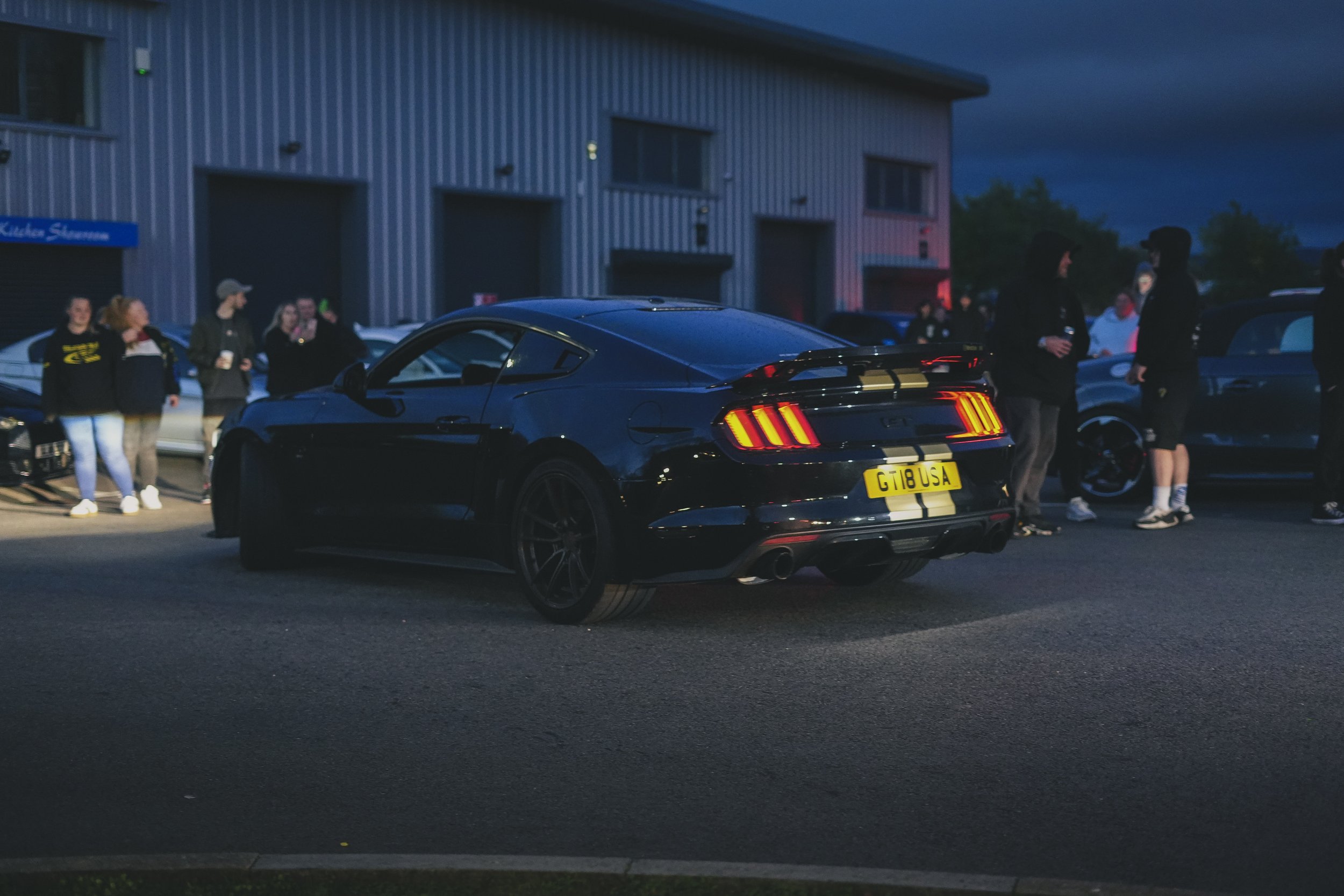 Black Ford Mustang with yellow racing stripes and a yellow license plate that reads GT 18 USA, parked outdoors during evening, with a group of people standing nearby in casual clothing, in front of a modern industrial building.