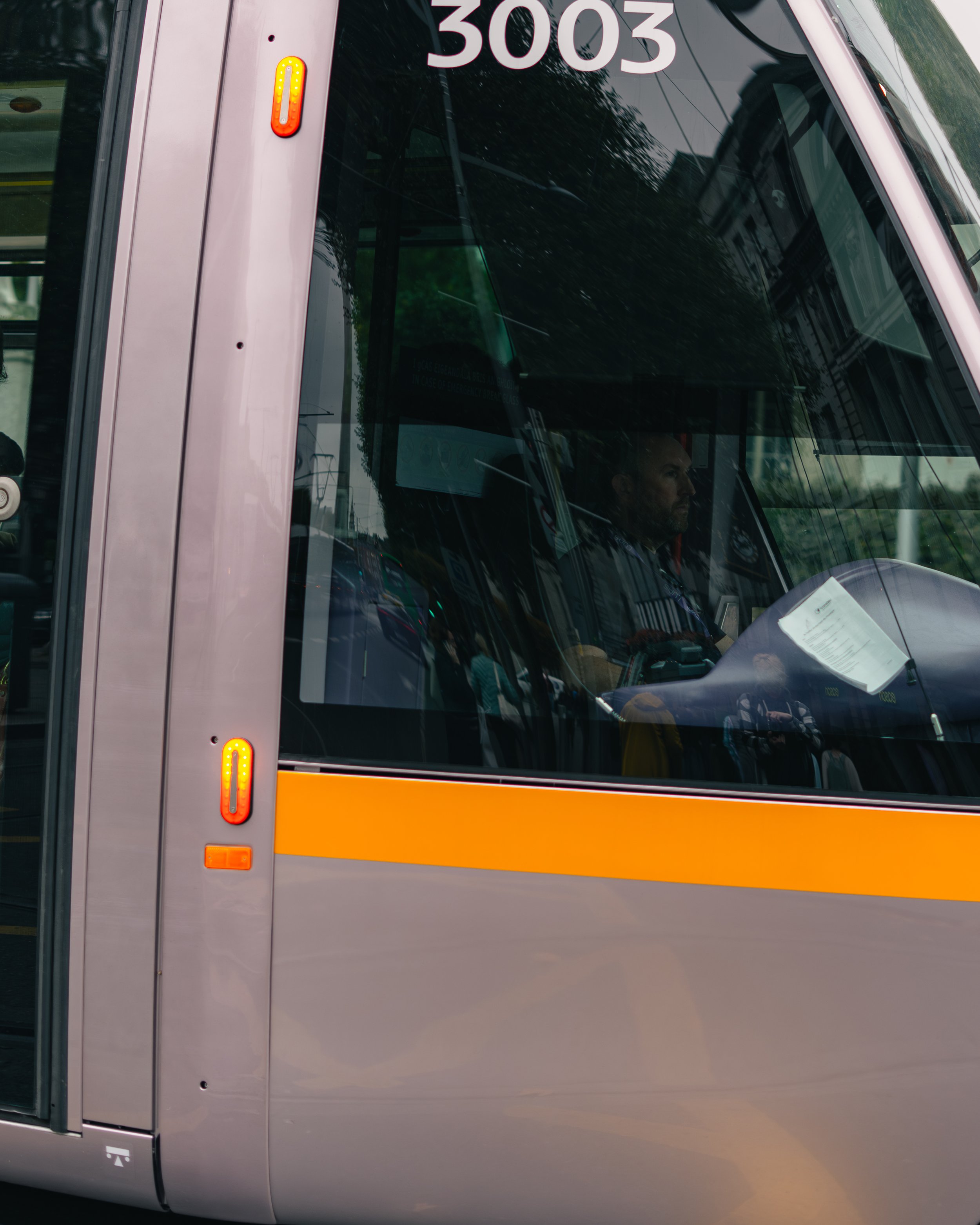 Close-up of a streetcar or light rail vehicle with the number 3003, showing the driver inside through the front windshield. The streetcar has a gray body with an orange stripe. Reflections of buildings and trees are visible on the glass.