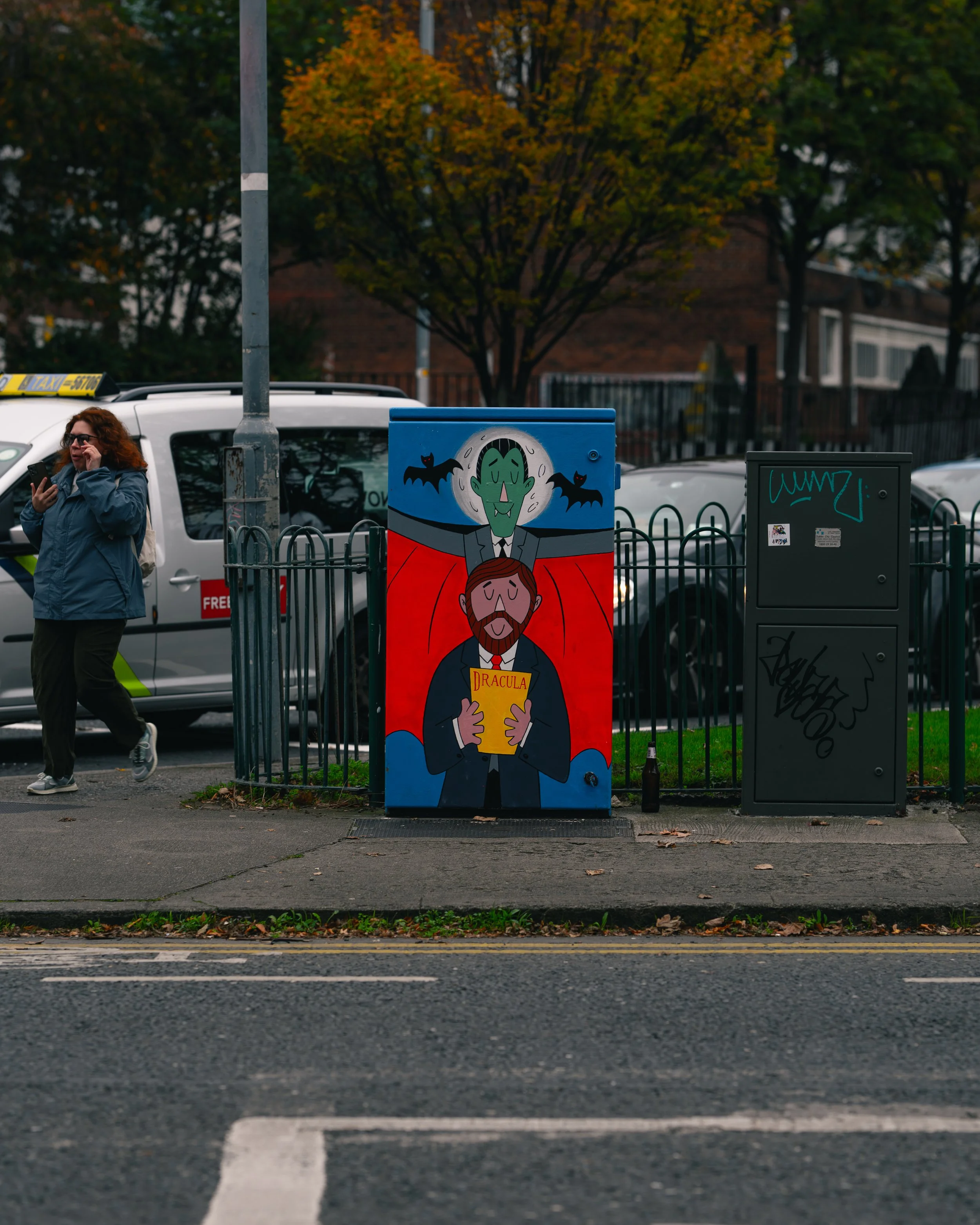 Street scene with colorful graffiti art of a vampire holding a book titled 'DRACULA' and a green face with bats behind it, a woman talking on a mobile phone walking past, parked cars, and trees with autumn leaves.
