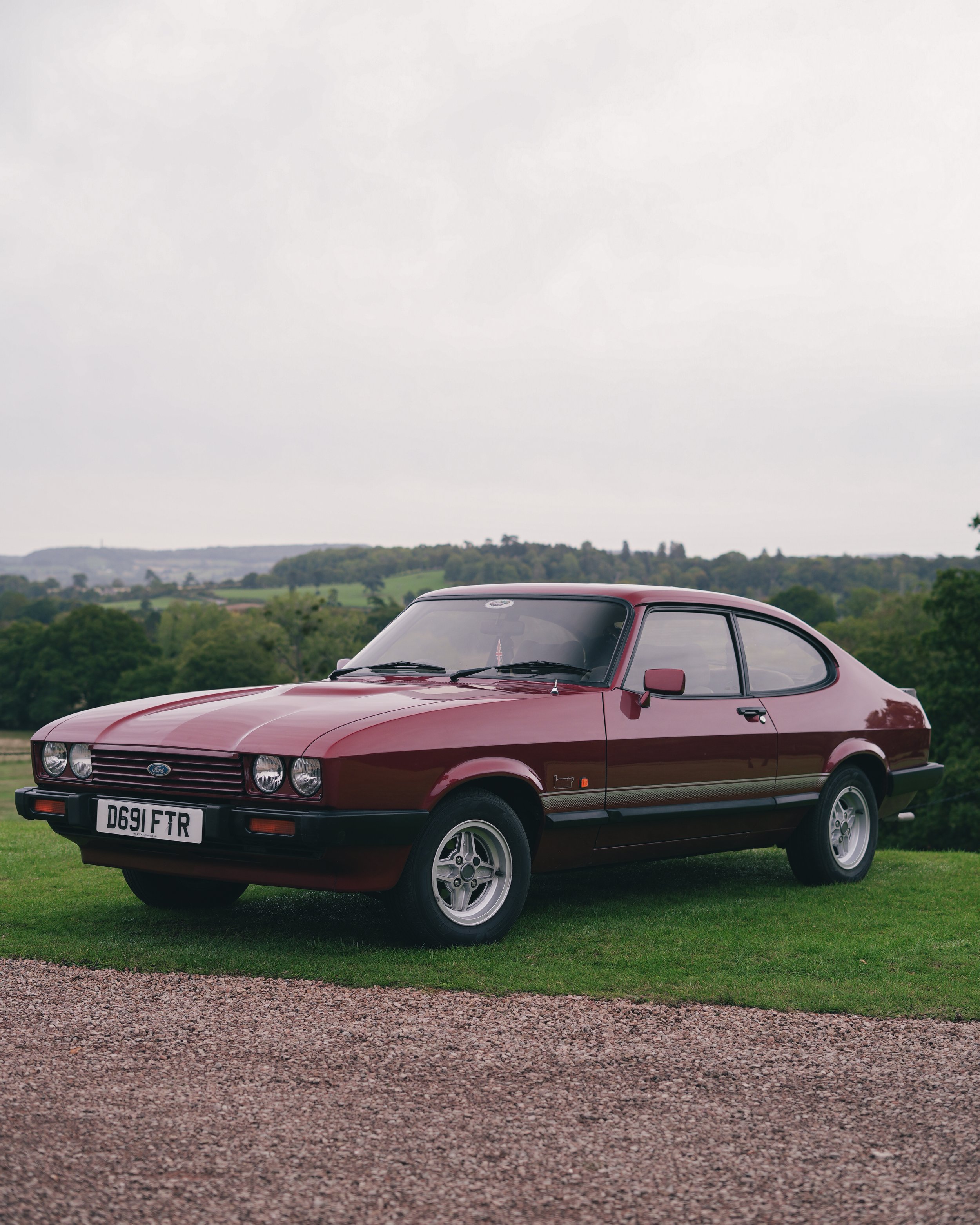 A vintage red Ford Capri car parked on a grassy area with rolling hills and trees in the background.