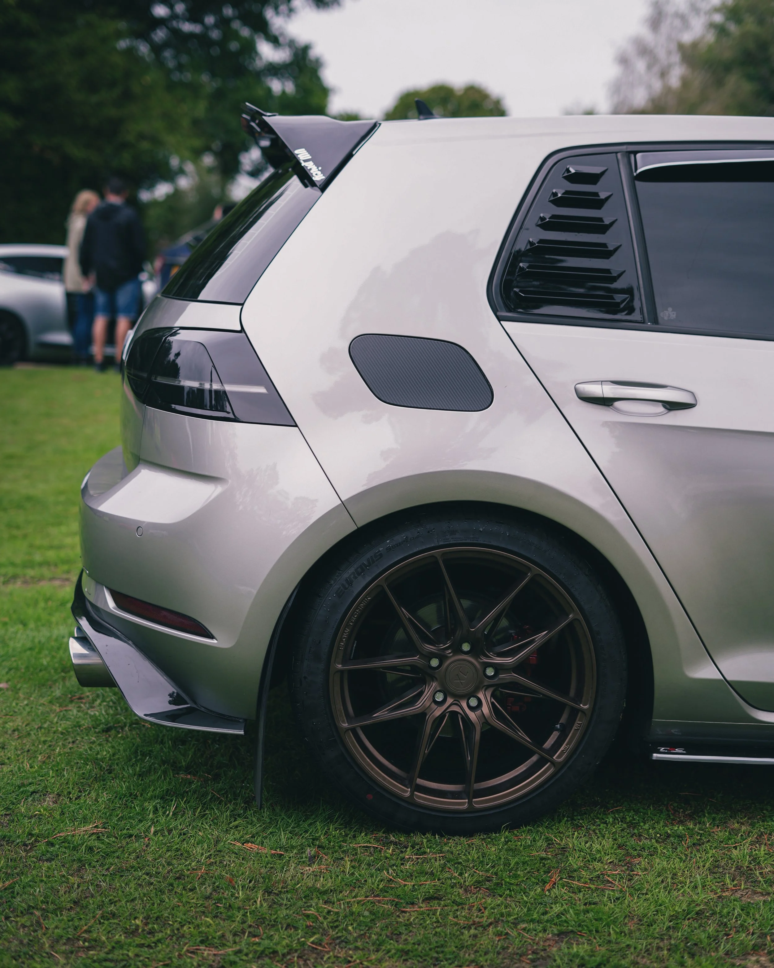 Close-up of the rear side of a silver sports car with black wheels, parked on grass during an outdoor car event, with people and trees in the background.