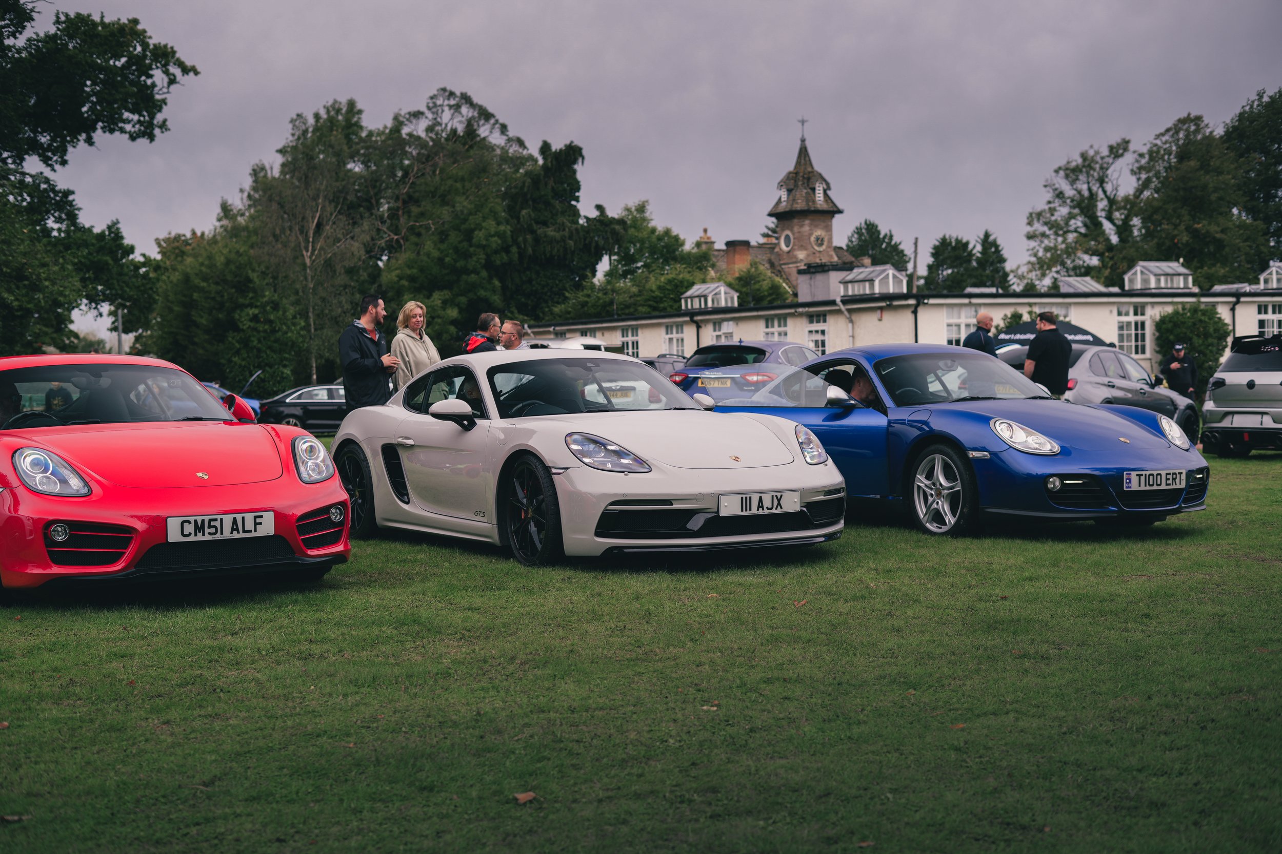 Three Porsche sports cars parked on a grassy field, with a group of people standing nearby and a building with a tower in the background.