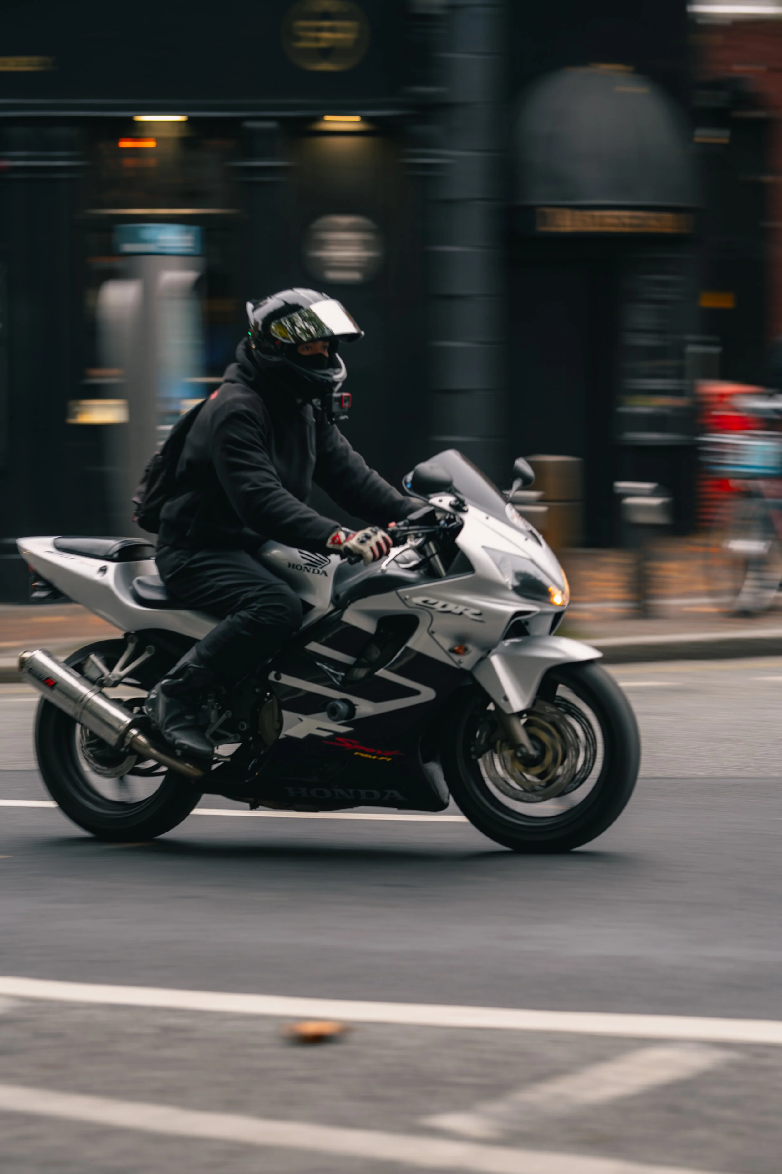 A motorcyclist dressed in black riding a Silver Honda sport bike on a city street.