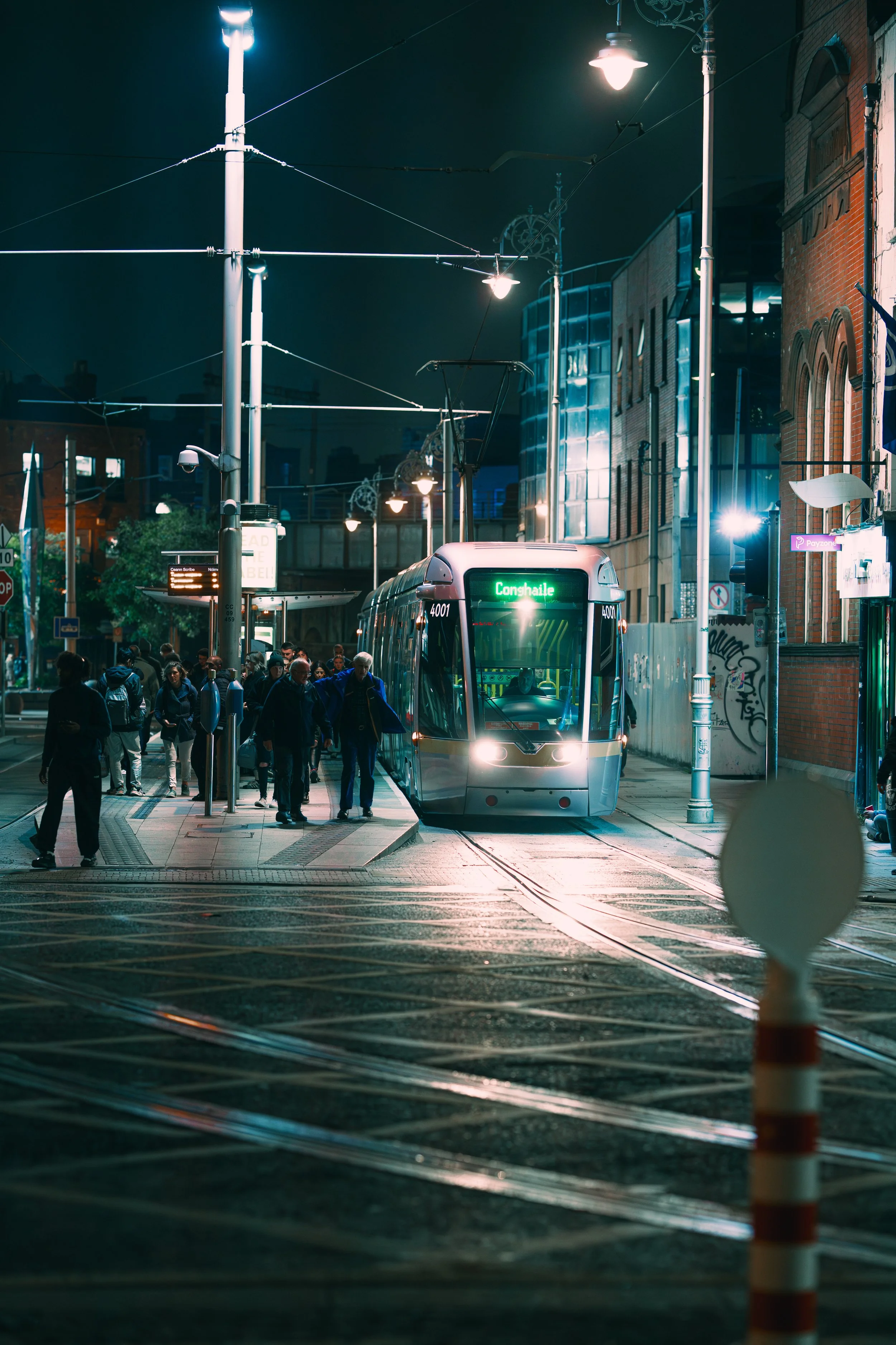Night scene of a modern tram stopped at a city tram stop with passengers boarding. The tram has a digital display showing its destination, 'Conxhaile.' Surrounding buildings are illuminated, and streetlights light up the scene.