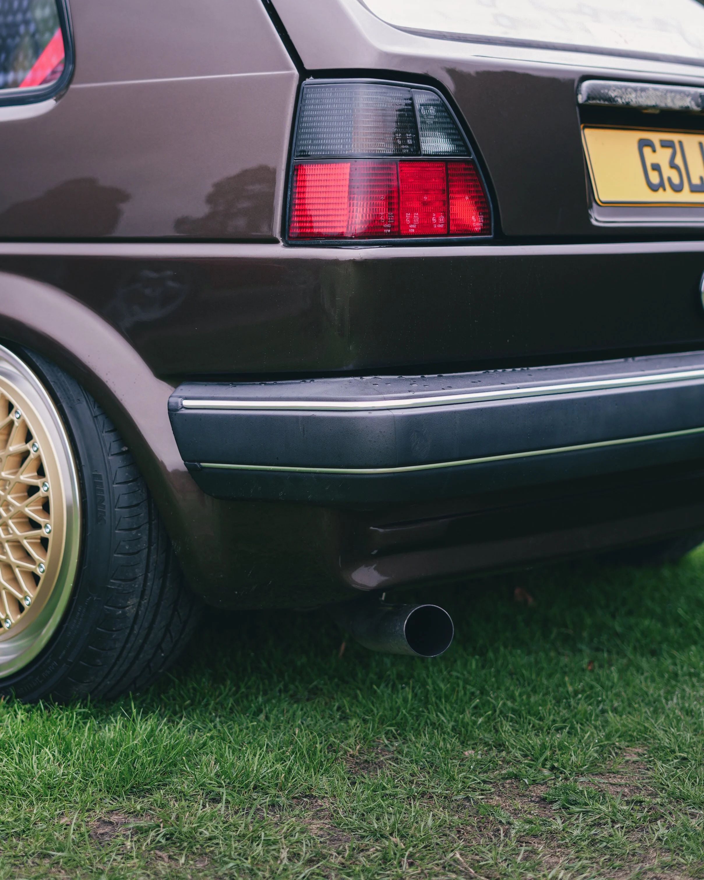 Close-up of the rear left side of a black vintage Volkswagen Golf car on grass, showing taillight, gold wheel with gold spokes, and exhaust pipe.