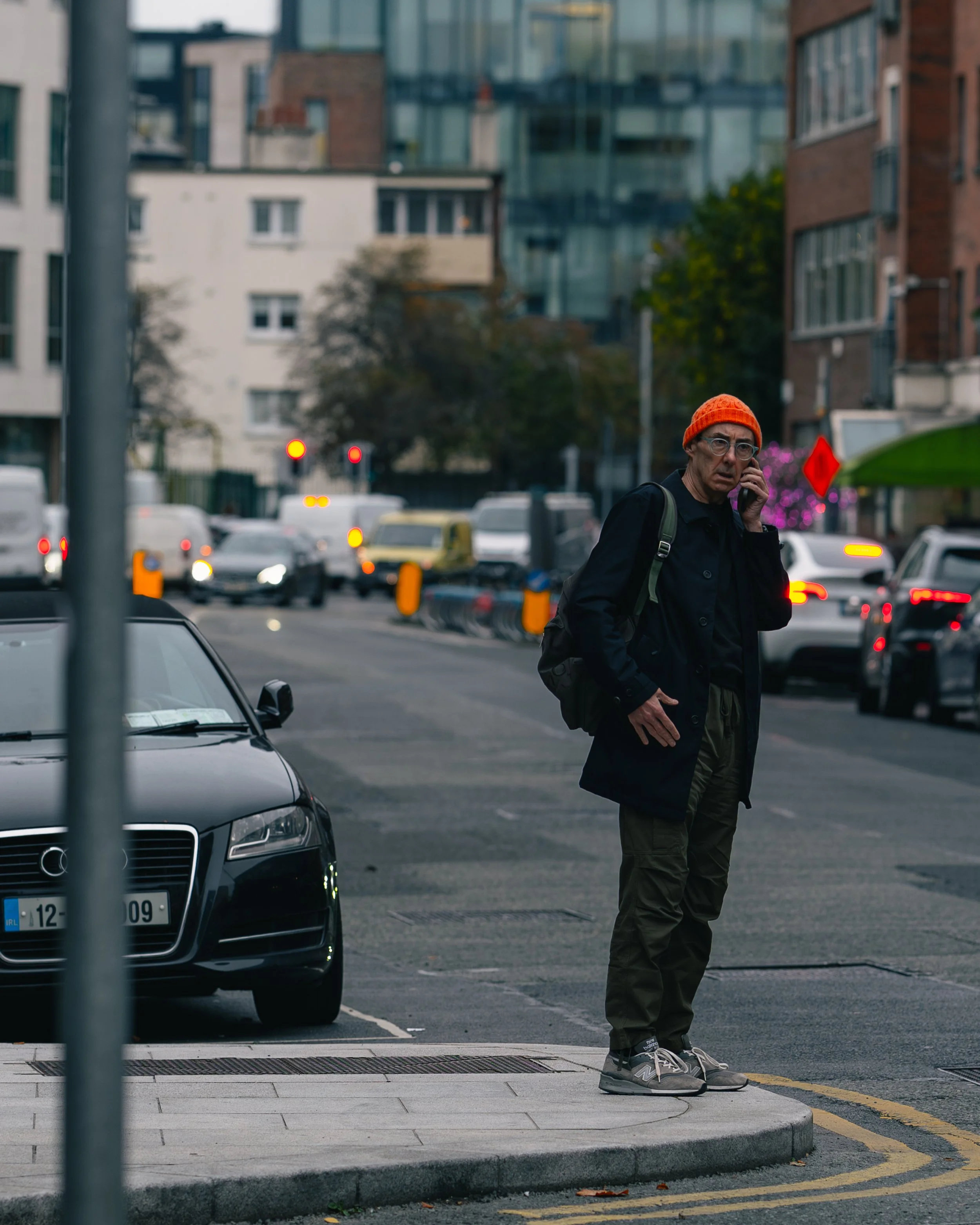 A man standing on a city sidewalk, talking on a mobile phone, wearing a bright orange hat, dark jacket, green pants, and sneakers, with traffic and buildings in the background.