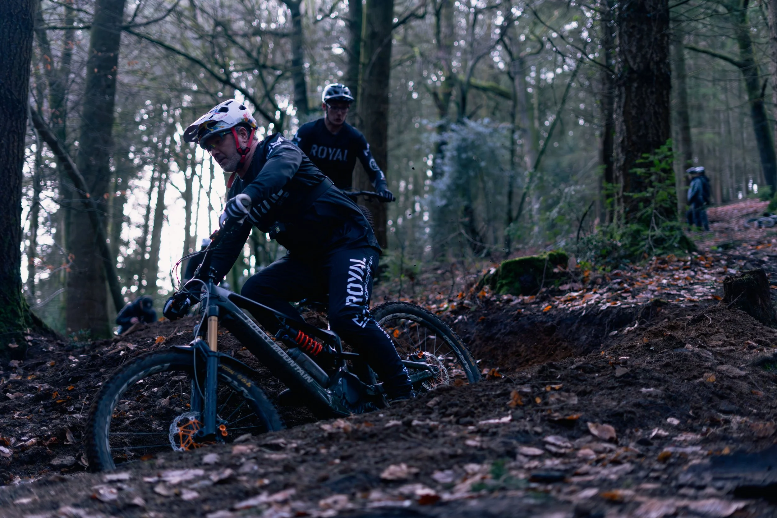 A group of mountain bikers riding down a dirt trail in a wooded forest with trees and fallen leaves.