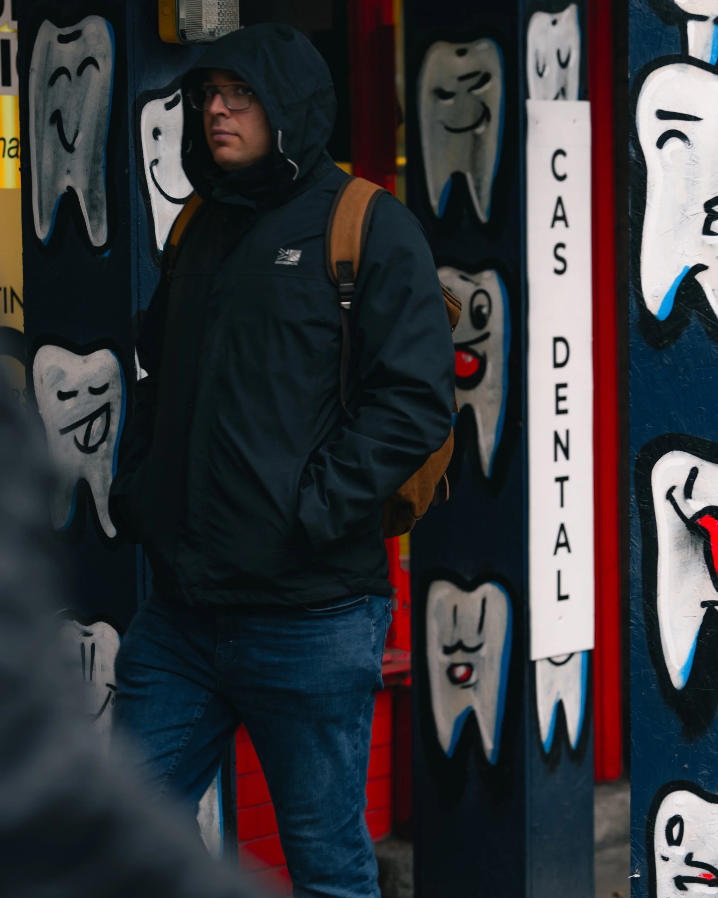 A man wearing a black hoodie, glasses, and a brown backpack standing against a wall decorated with cartoonish smiling teeth art, part of a dental clinic advertisement.