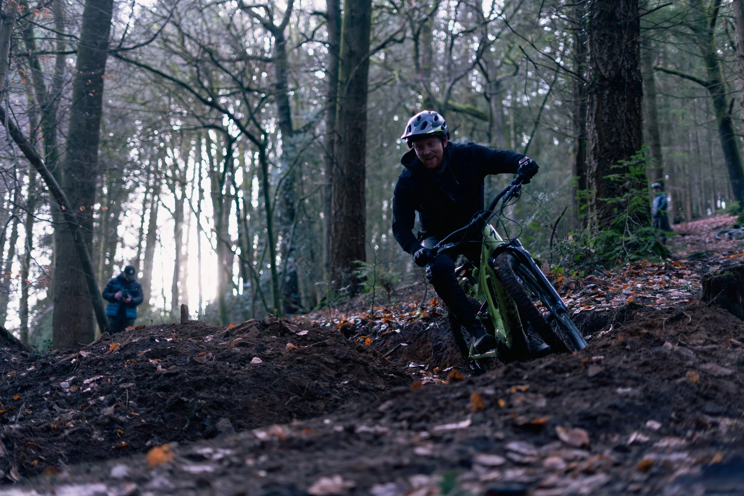 A person mountain biking on a dirt trail in a forest, wearing a helmet and black clothing, with other people visible in the background.