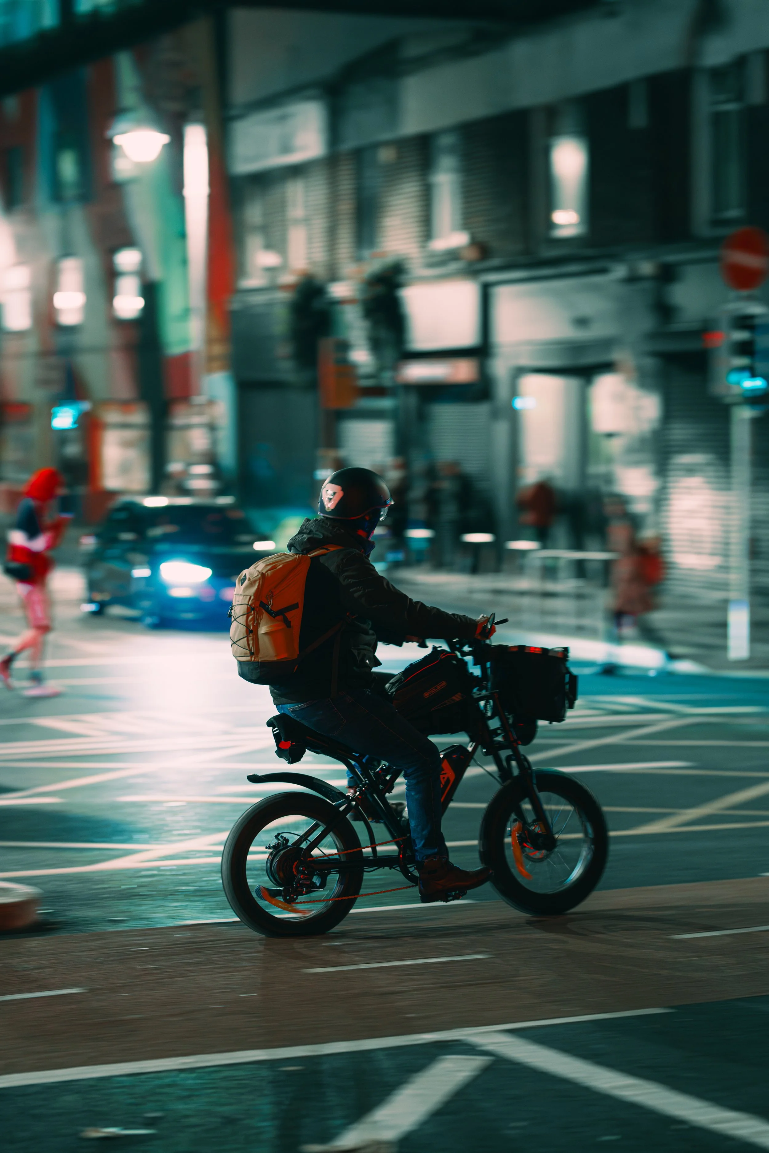 A person riding an electric bicycle through a city street at night, with other pedestrians and cars in the background.