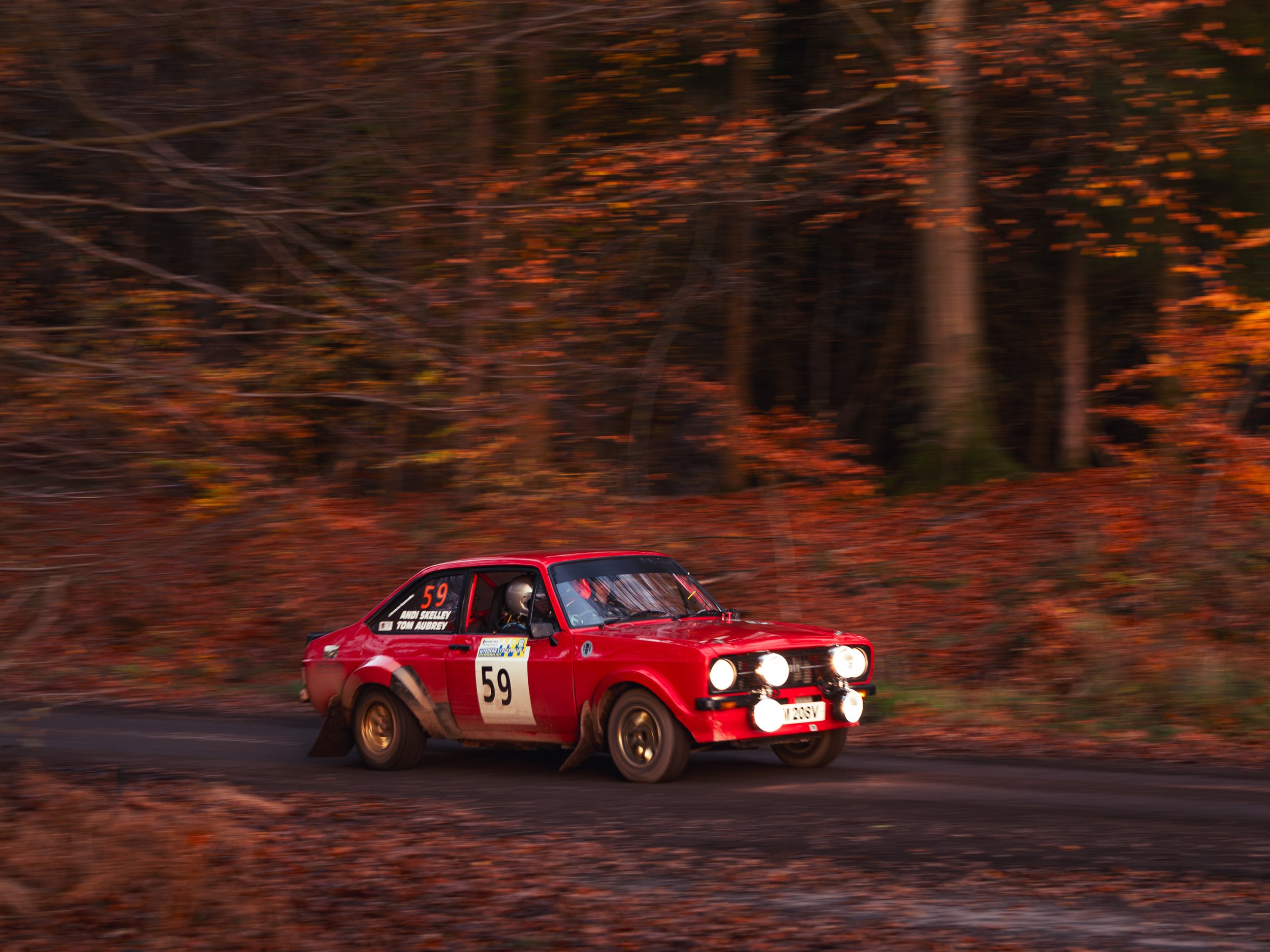 A red rally car with the number 59 racing through an autumn forest, with orange and brown leaves falling around it. wye dean rally 