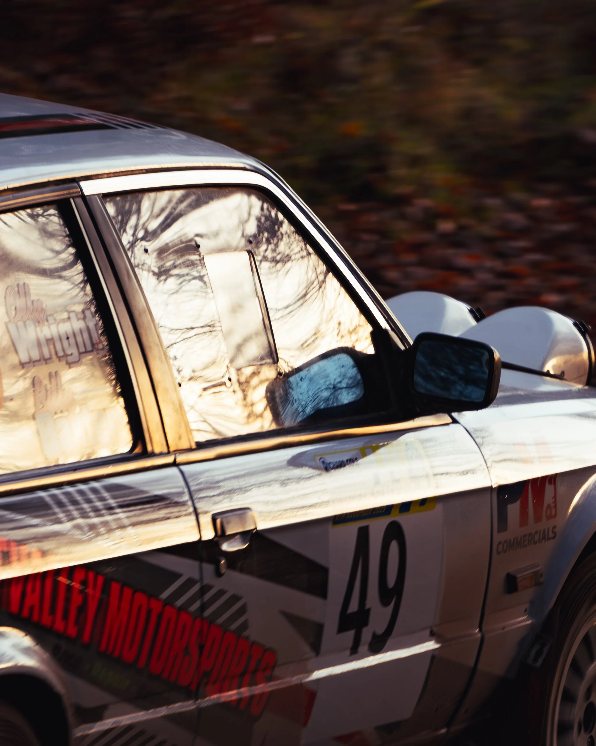 Close-up of a vintage race car with the number 49, reflecting trees in the windshield and part of a sign reading 'Valley Motorsports.' wye dean rally 