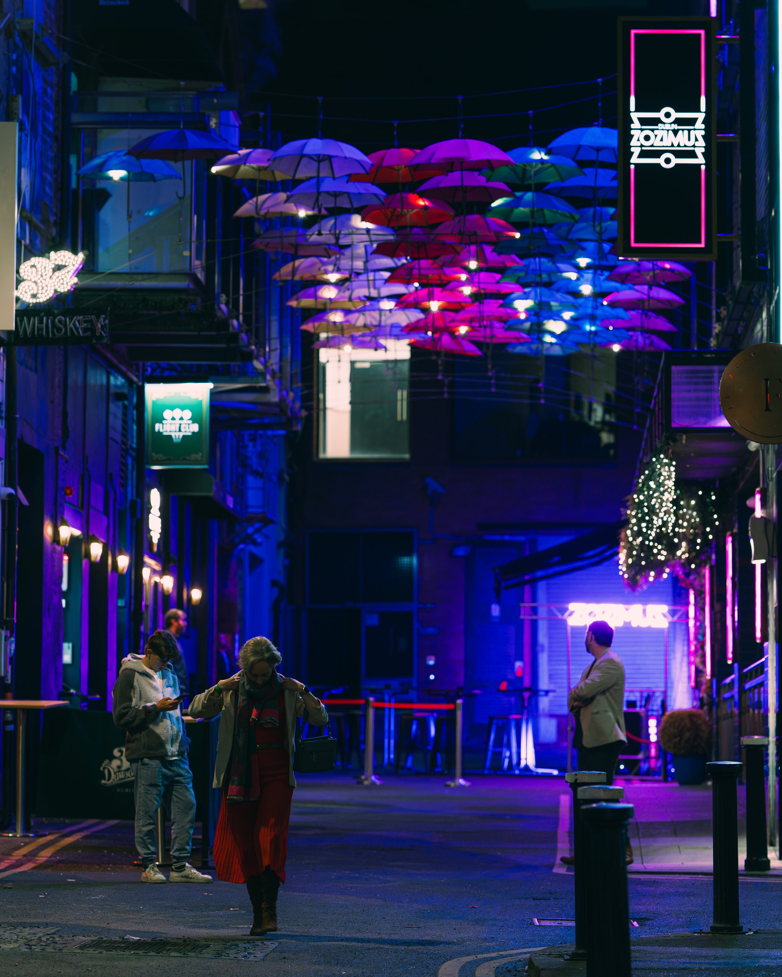 Night scene of a city street decorated with colorful umbrellas hanging overhead and neon signs. A few people are walking, and the street is illuminated with blue and purple lighting.