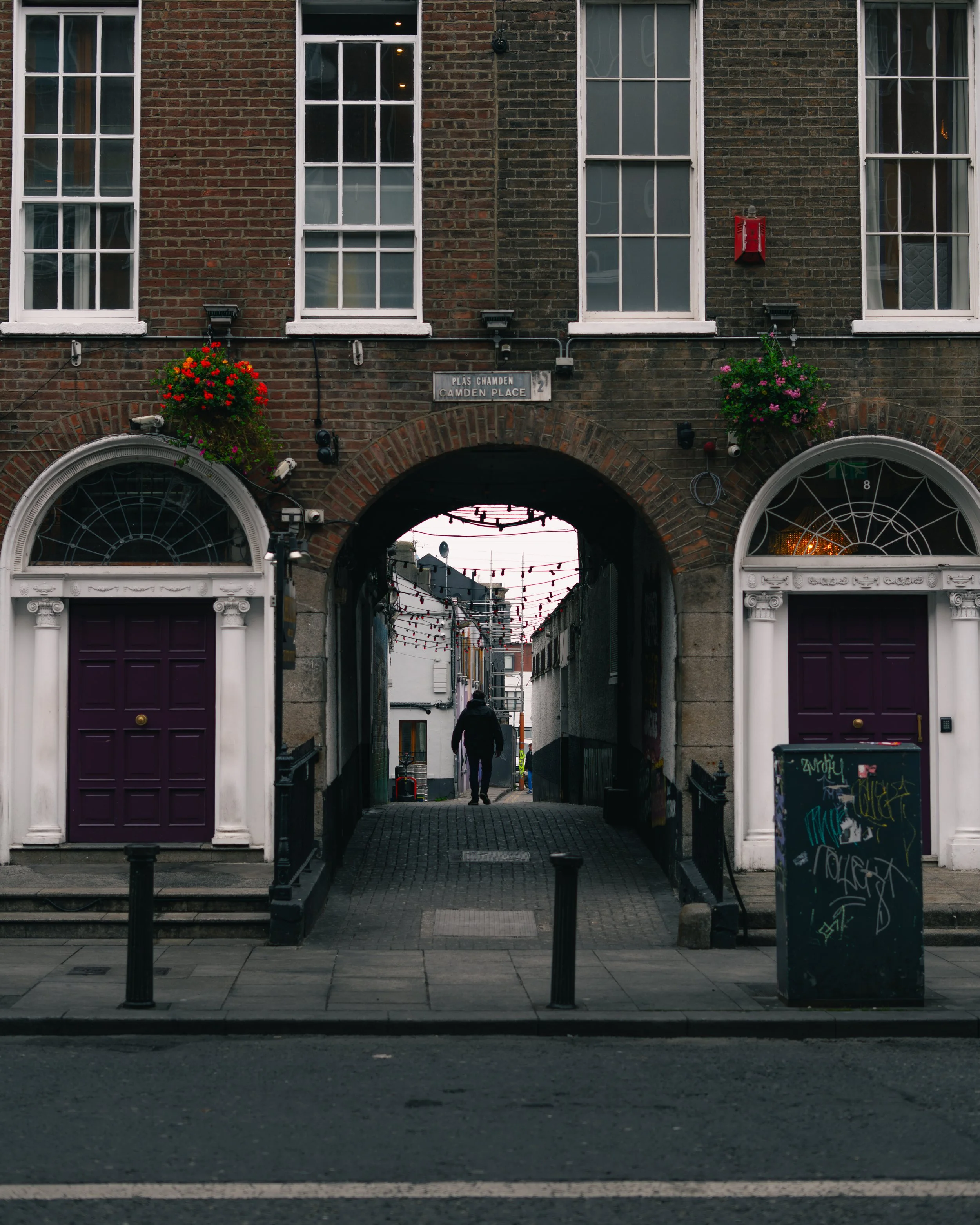 An arched entrance in a brick building with white-framed windows and purple doors, leading to a narrow alleyway decorated with string lights, with a person walking away in the distance.