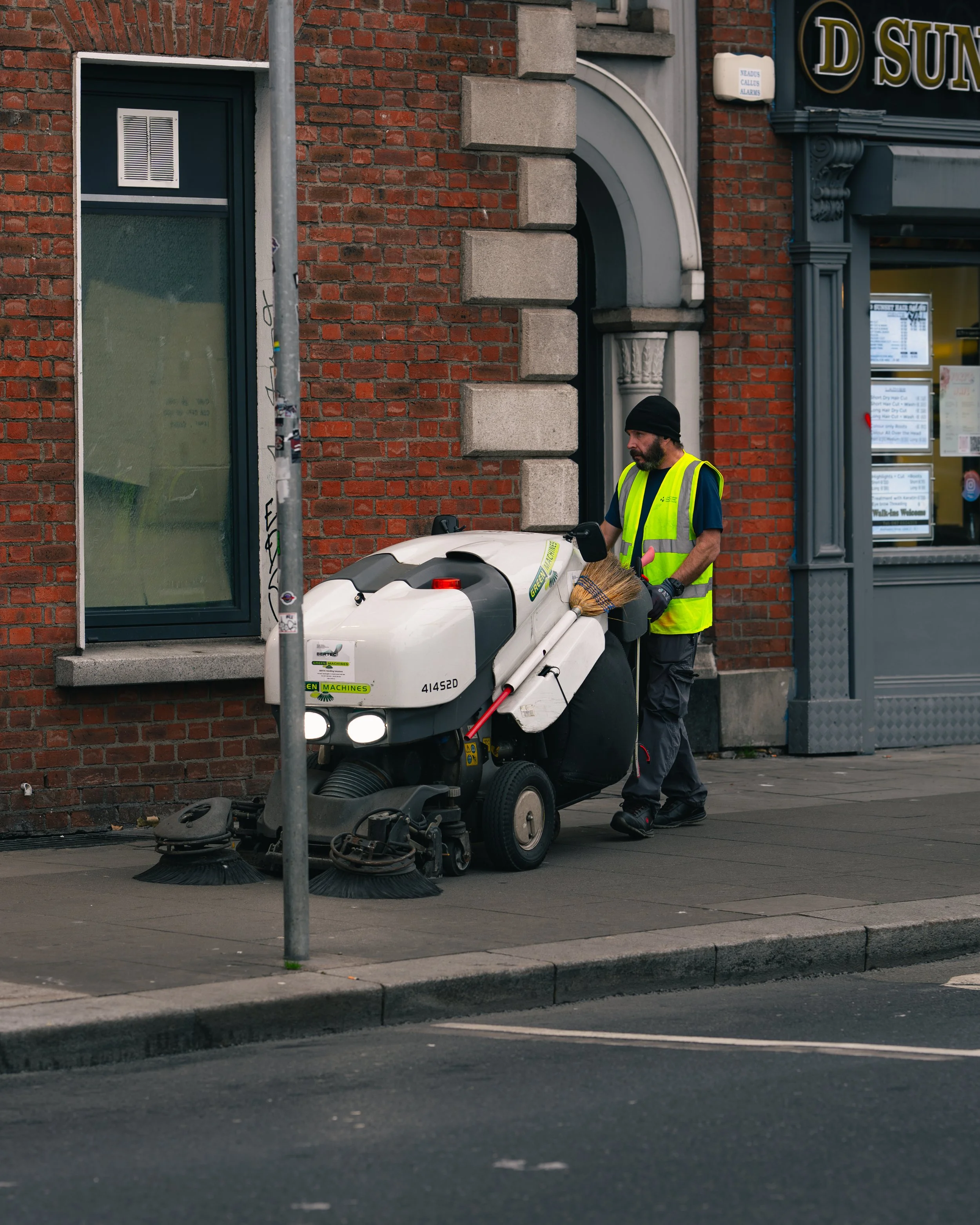 A street cleaner in a yellow safety vest and black beanie using a street sweeping machine on the sidewalk in front of a brick building.