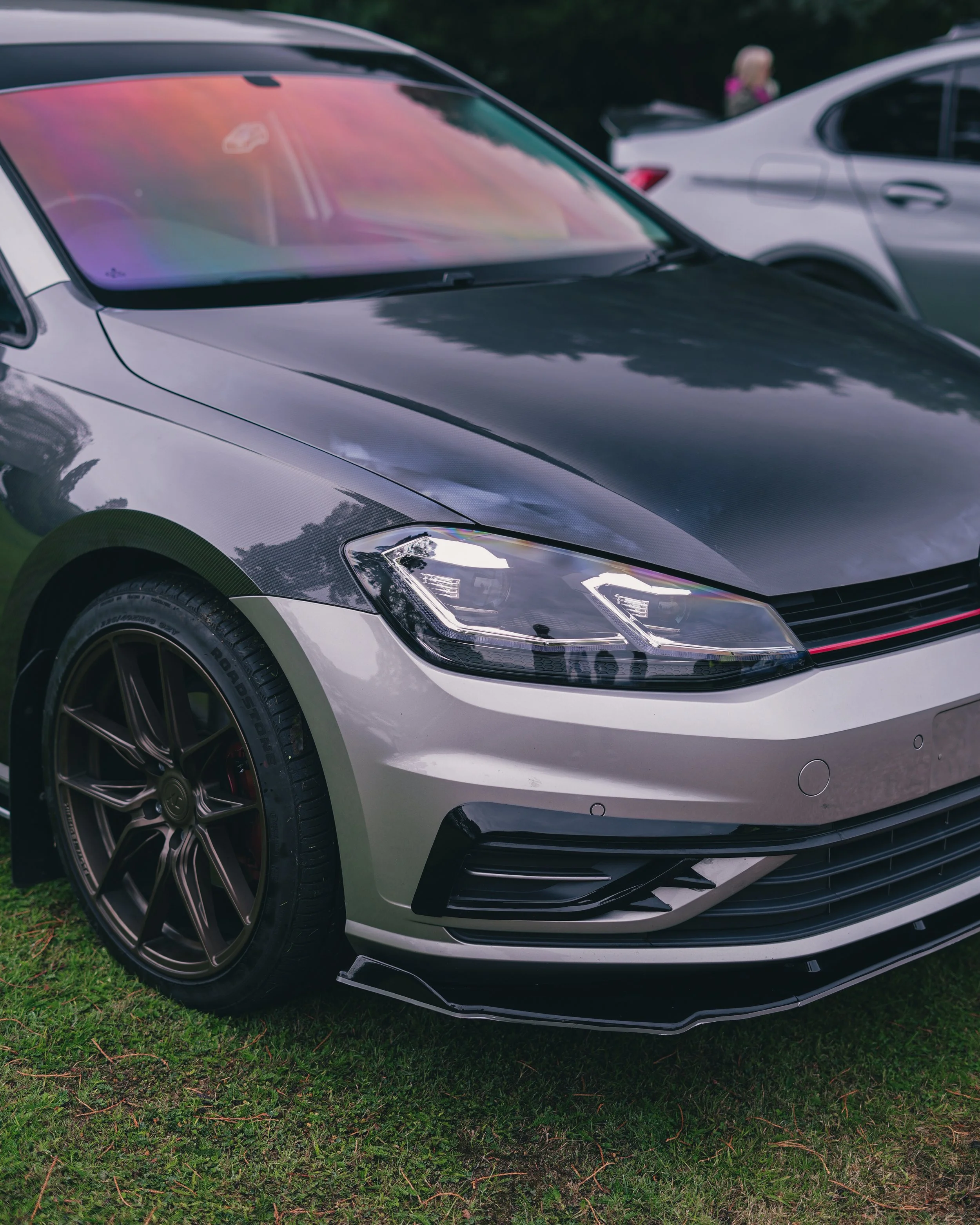 Close-up of a black and silver modern sedan parked on grass, with other vehicles and people visible in the background.