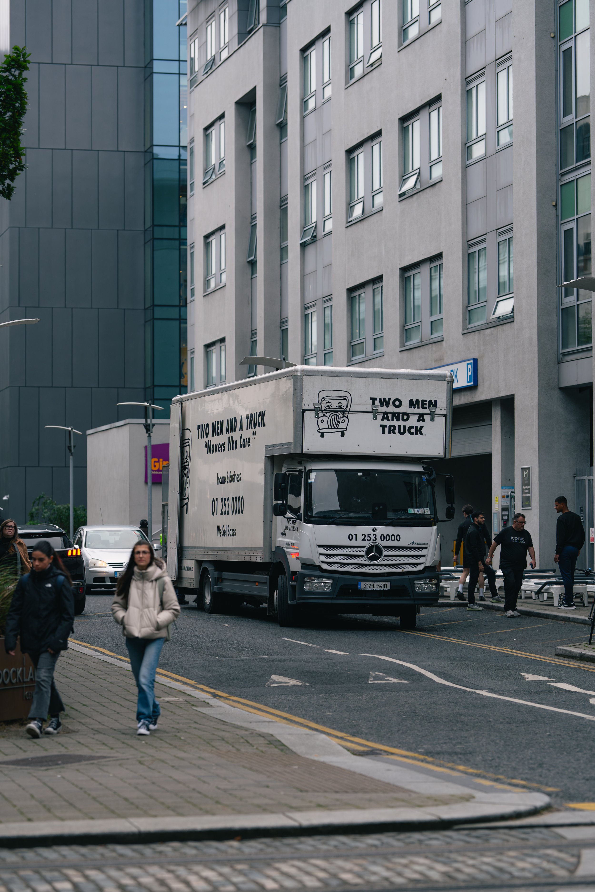 A street scene featuring a moving truck with the company name 'Two Men and a Truck' and several pedestrians walking on the sidewalk in an urban area with modern high-rise buildings.