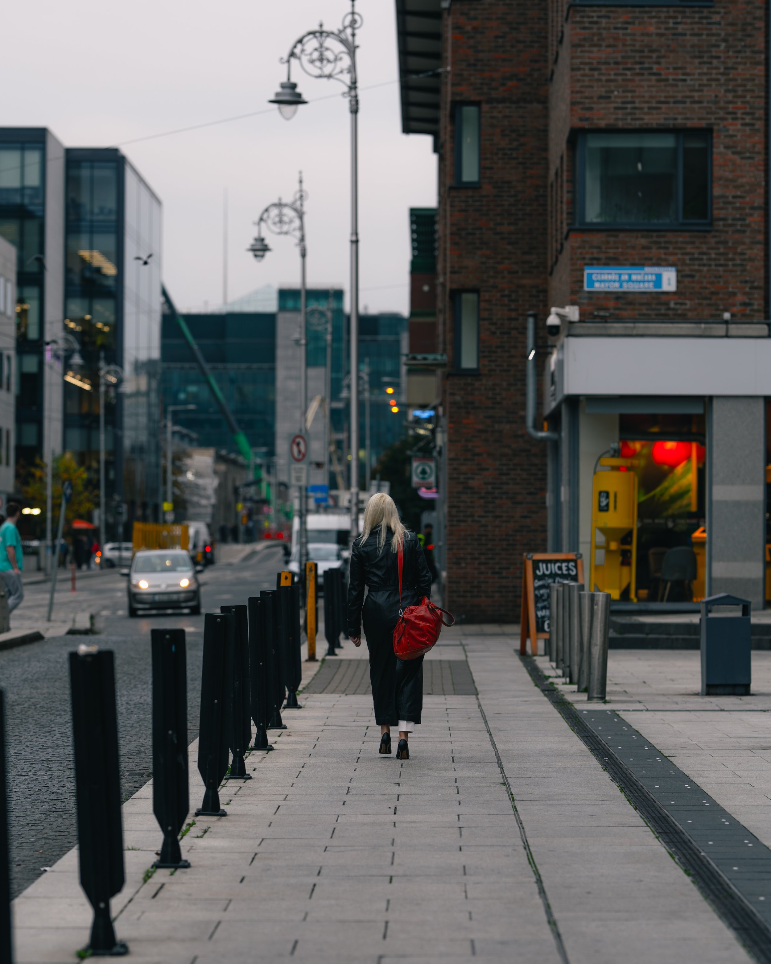 A woman with blonde hair, wearing a black coat and black shoes, walking alone on a city sidewalk carrying a large red bag. The background includes modern glass buildings, brick buildings, street lamps, and a storefront with a sign for juices.