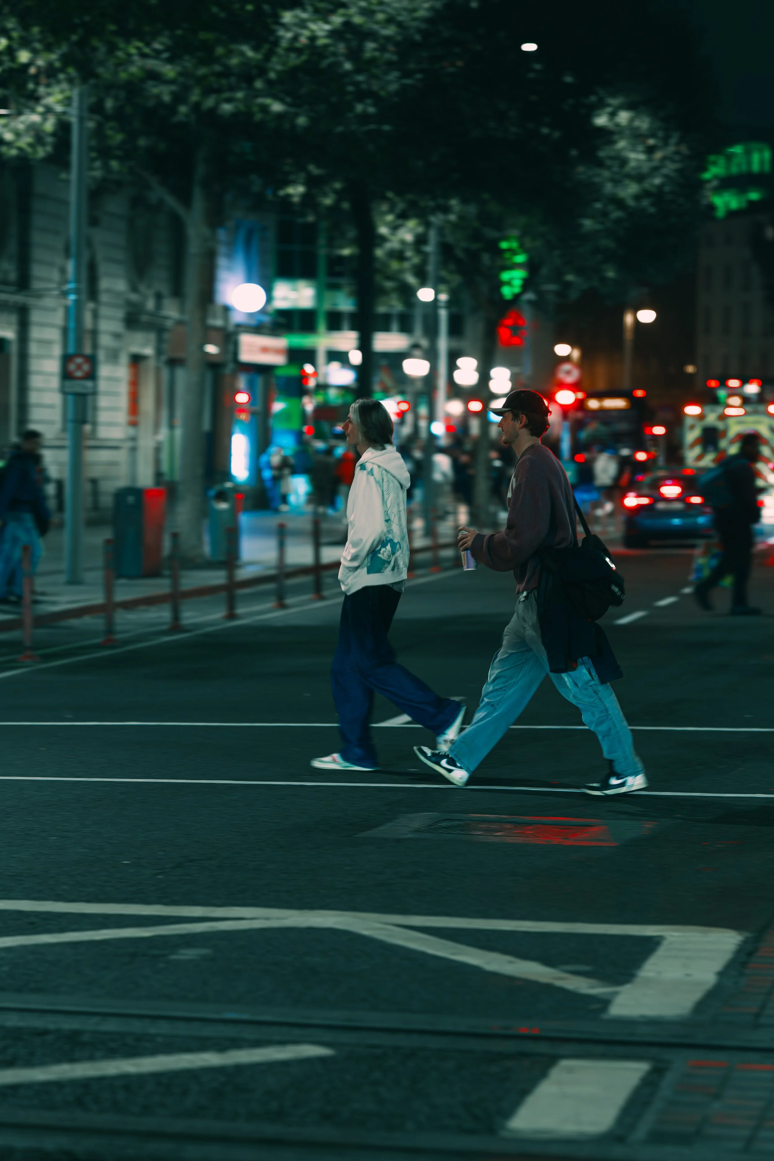Two people crossing a city street at night, with traffic and illuminated signs in the background.