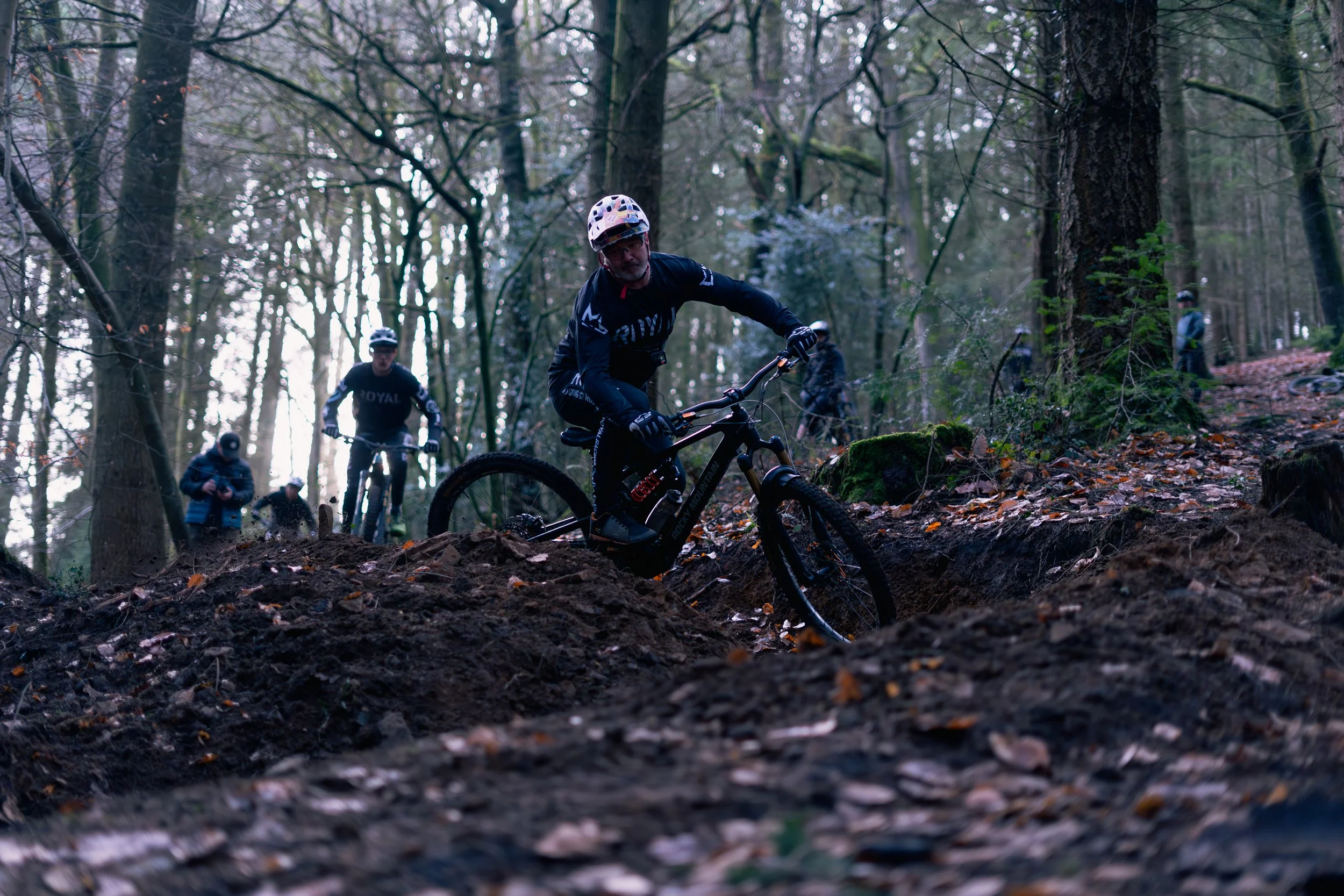A group of mountain bikers riding on a dirt trail through a forest with tall trees and fallen leaves on the ground