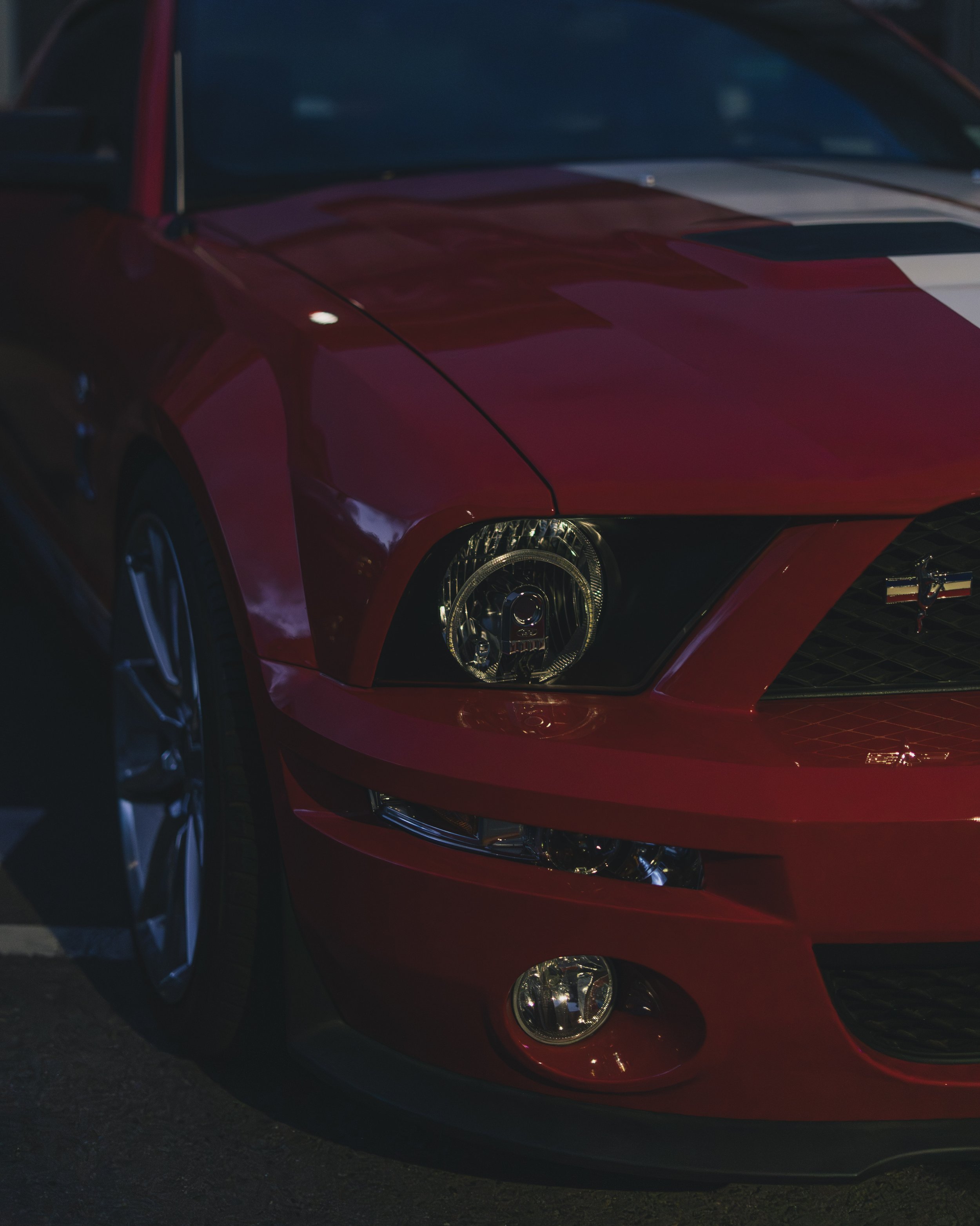 Close-up of a red Ford Mustang sports car showing the front left side, headlight, fog light, and part of the wheel with a dark background.