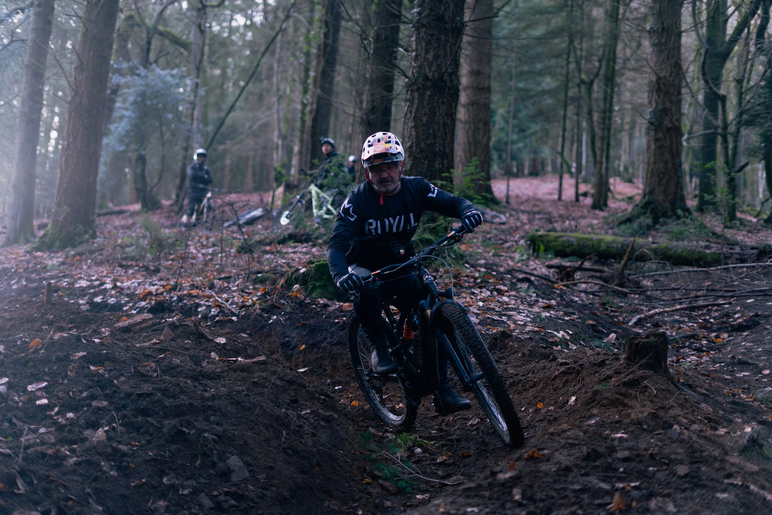 A person mountain biking on a rugged trail through a forest with two other bikers in the background.