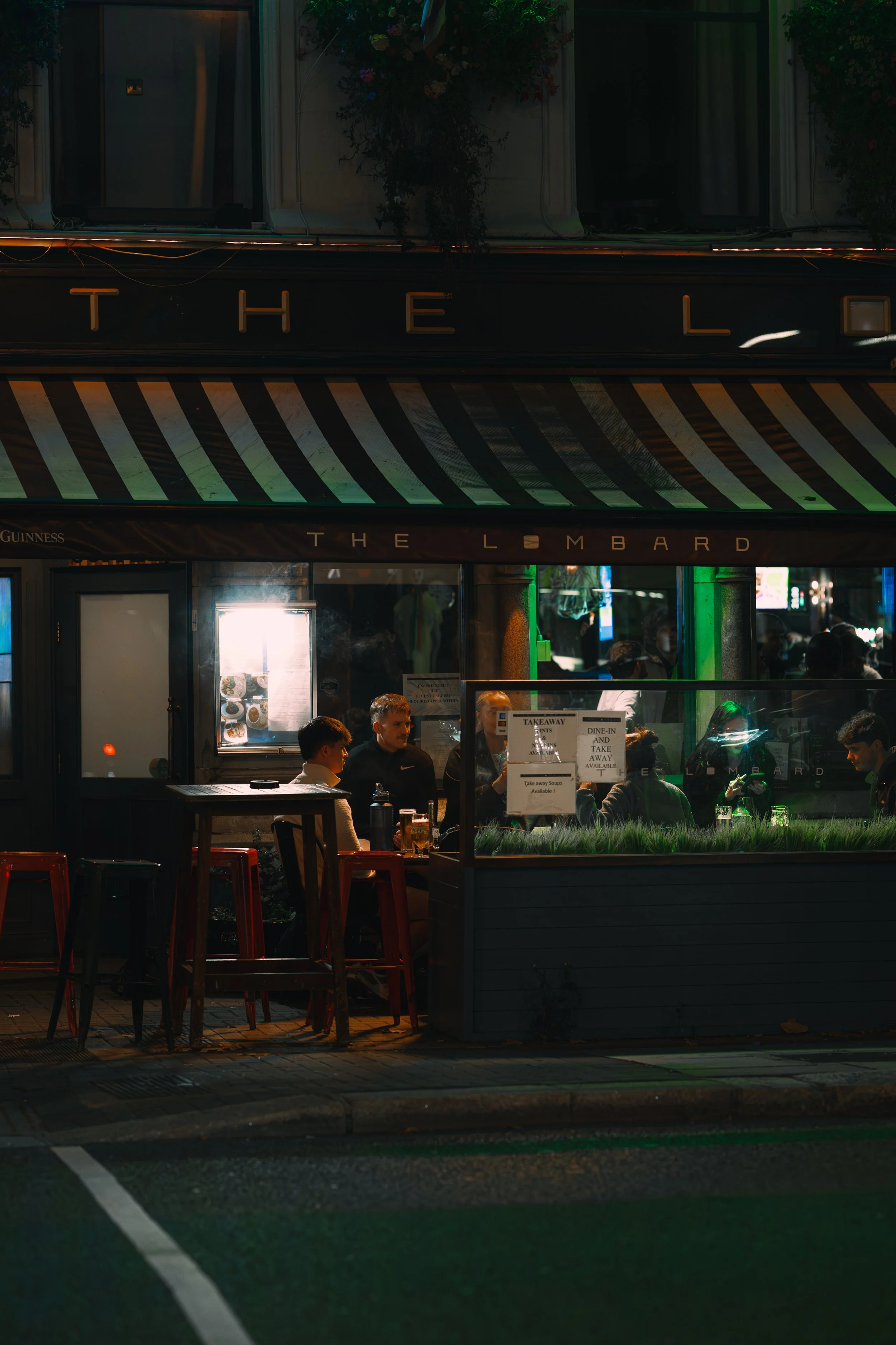 Nighttime scene of a pub with people sitting inside and outside, illuminated by lights and surrounded by dark street.