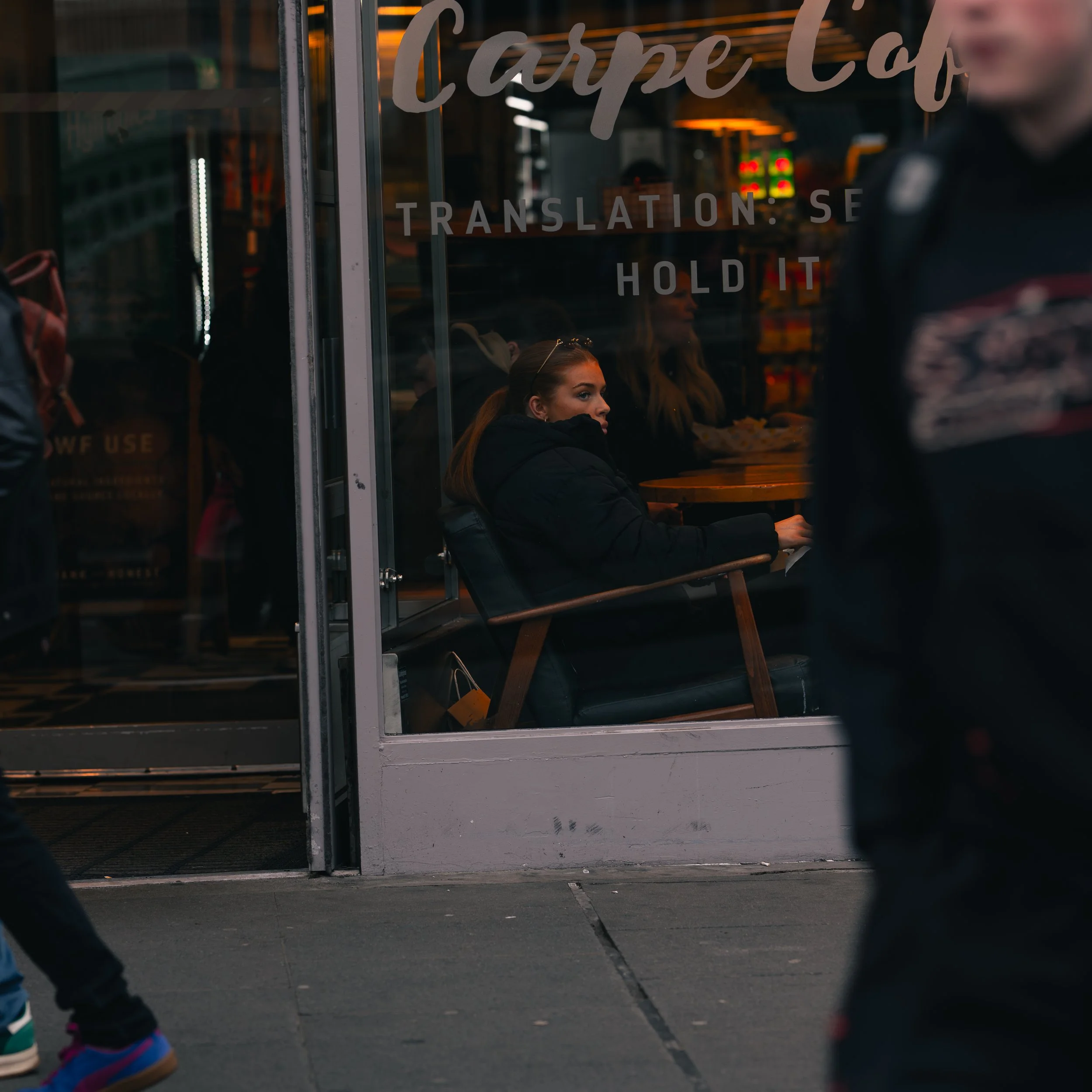 A woman sitting alone inside a café with a window, looking outside, with people walking by on the street. The café has a sign on the window that reads ‘Carpe Café TRANSLATION SE HOLD IT’.