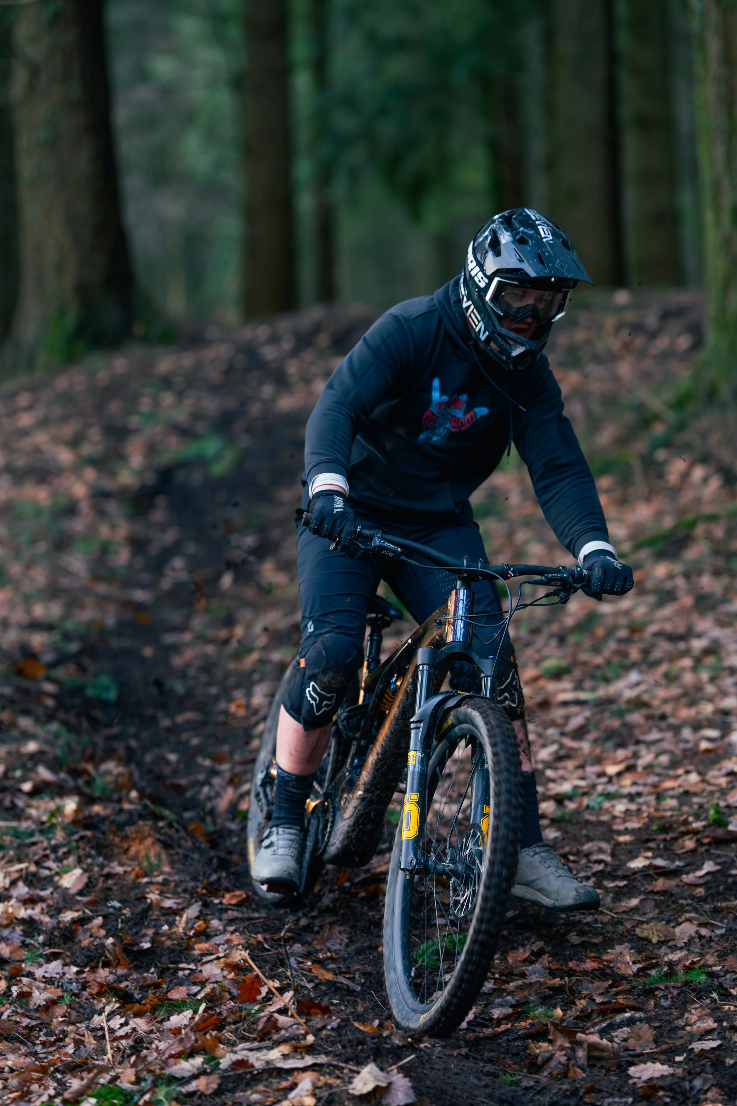 A person mountain biking on a wooded trail, wearing a helmet, gloves, and protective gear.