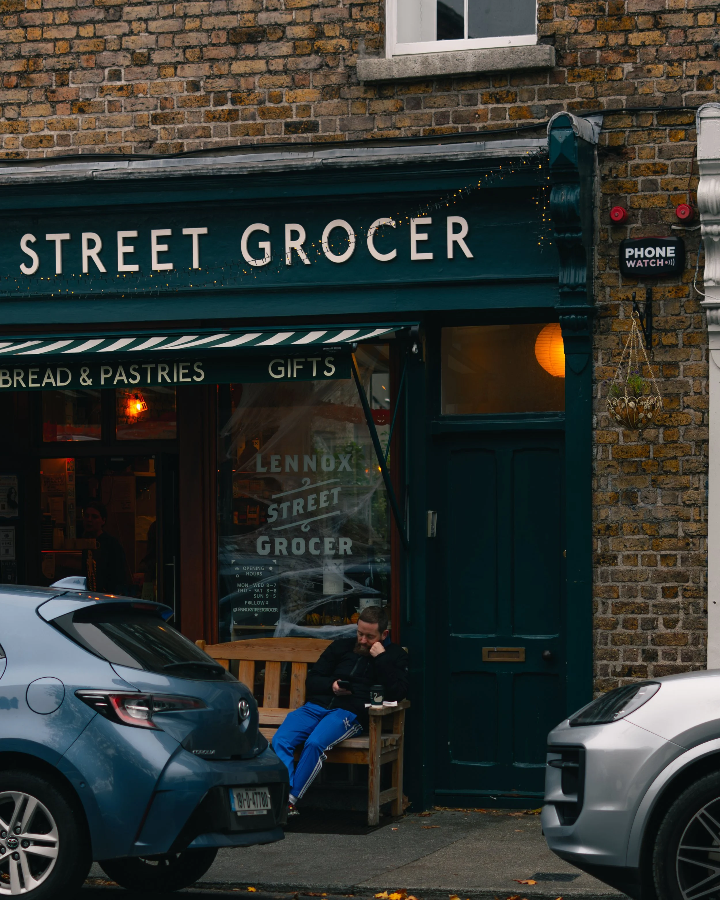 A man sitting on a wooden bench outside a bakery and pastry shop called Lennox Street Grocer. The shop has a black awning with white lettering, and a sign in the window with the shop's name. There are two cars parked nearby, one blue and one silver. 