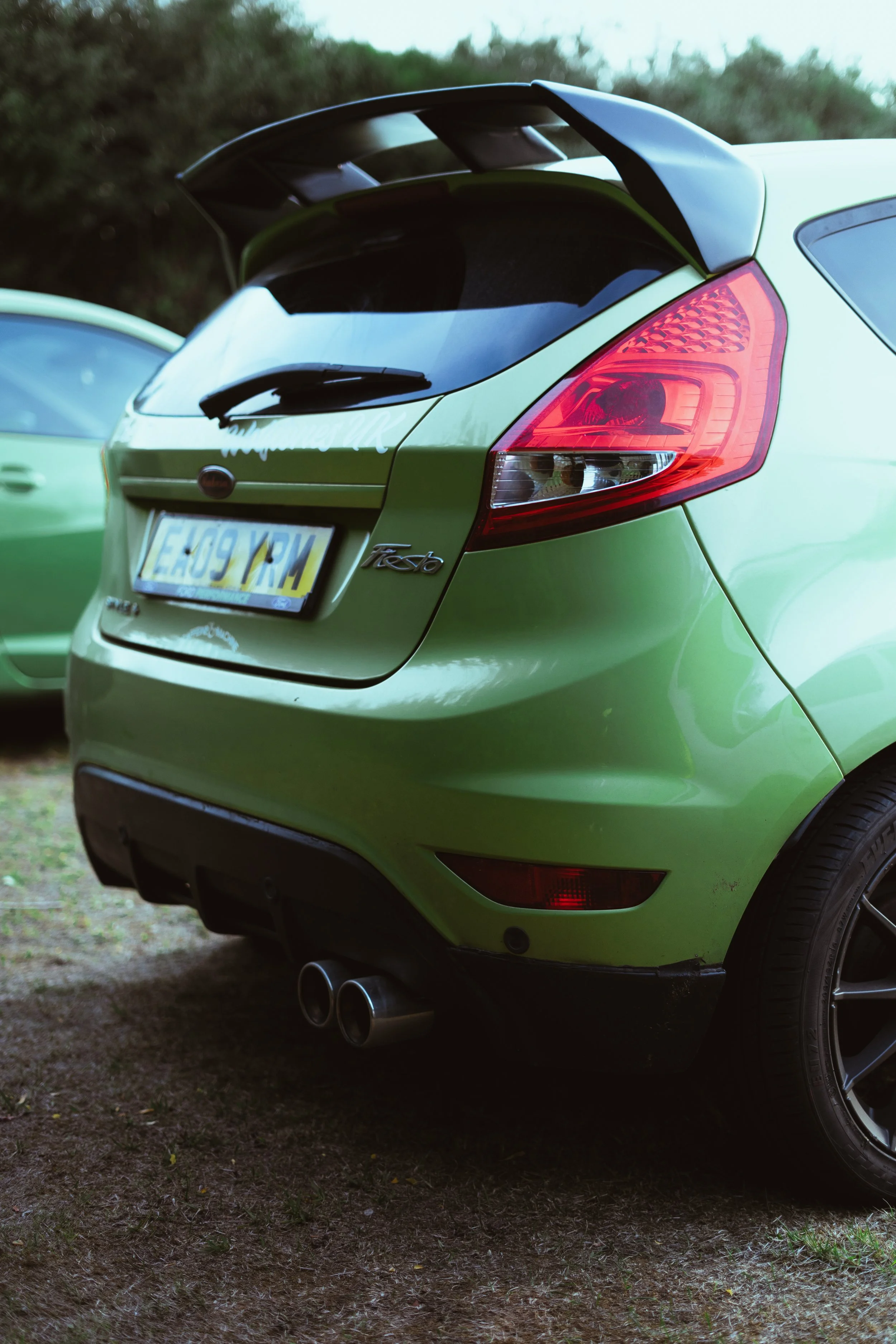 Bright green Ford Festa hatchback car parked at a car meet with a black rear spoiler and dual exhaust pipes visible at the rear.