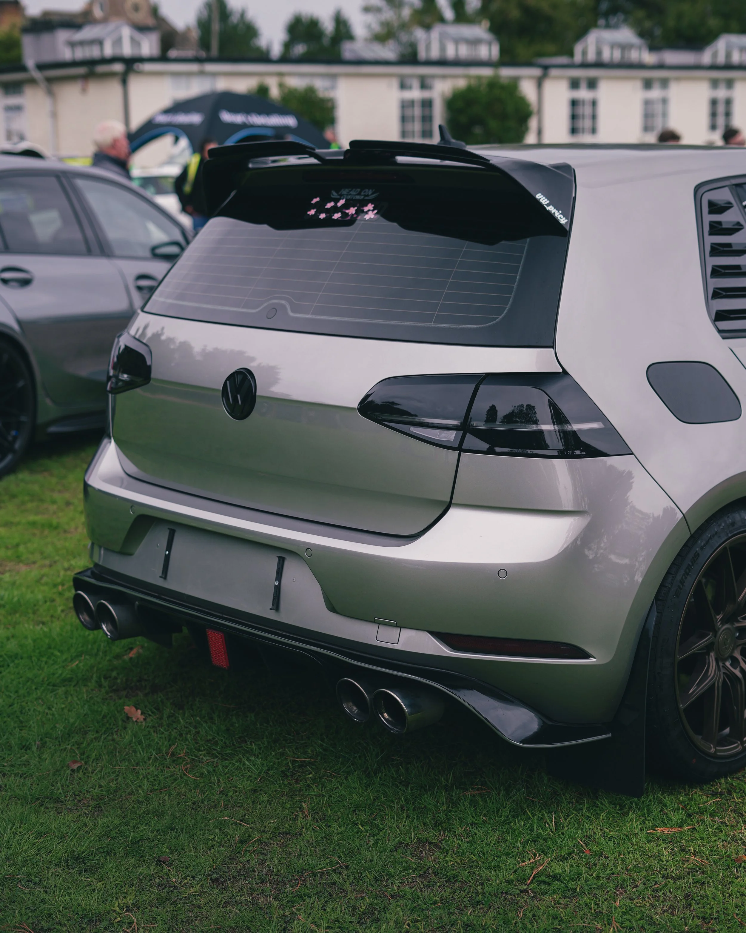 A silver Volkswagen car with black accents and quad exhaust pipes parked on a grassy area at an outdoor car show.