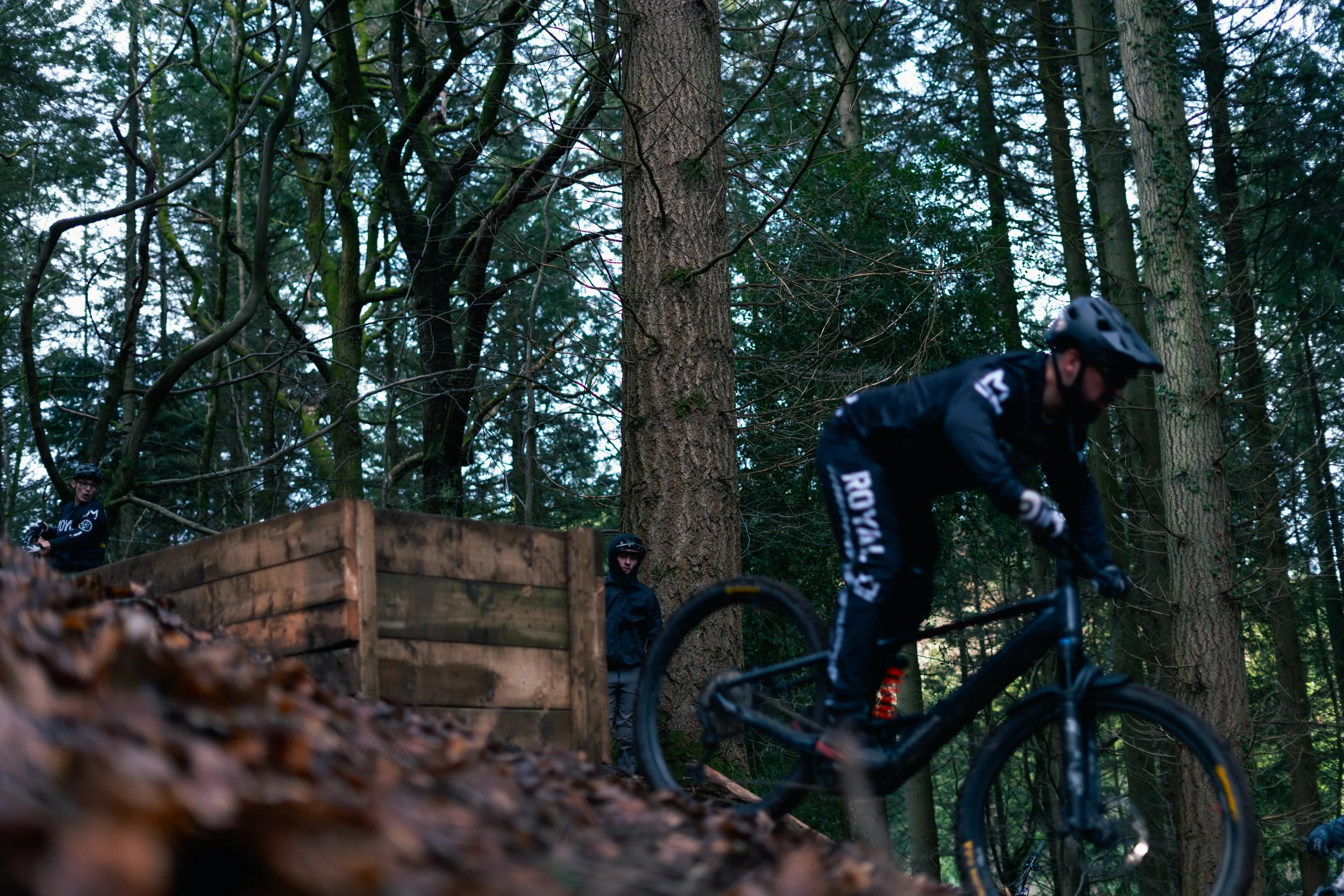 A mountain biker wearing a helmet riding downhill on a forest trail surrounded by tall trees, with two people on the side, one looking on and the other behind a wooden platform.