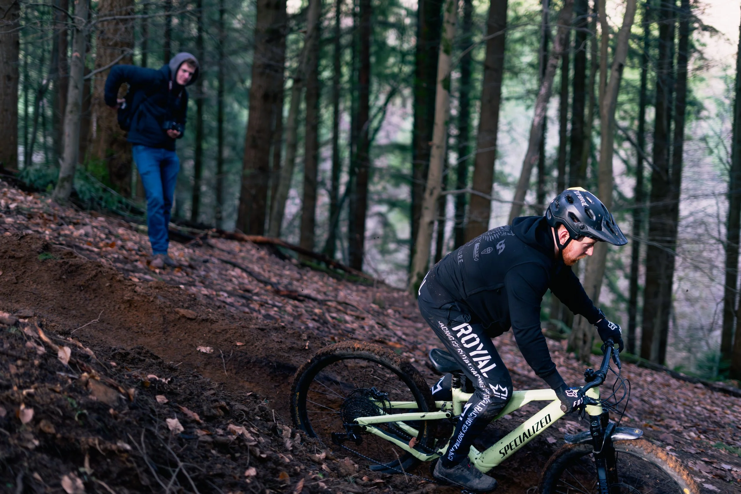 A mountain biker wearing a black helmet and black gear riding a yellow Specialized bike downhill on a forest trail, with a person in a black hoodie and blue jeans walking nearby.