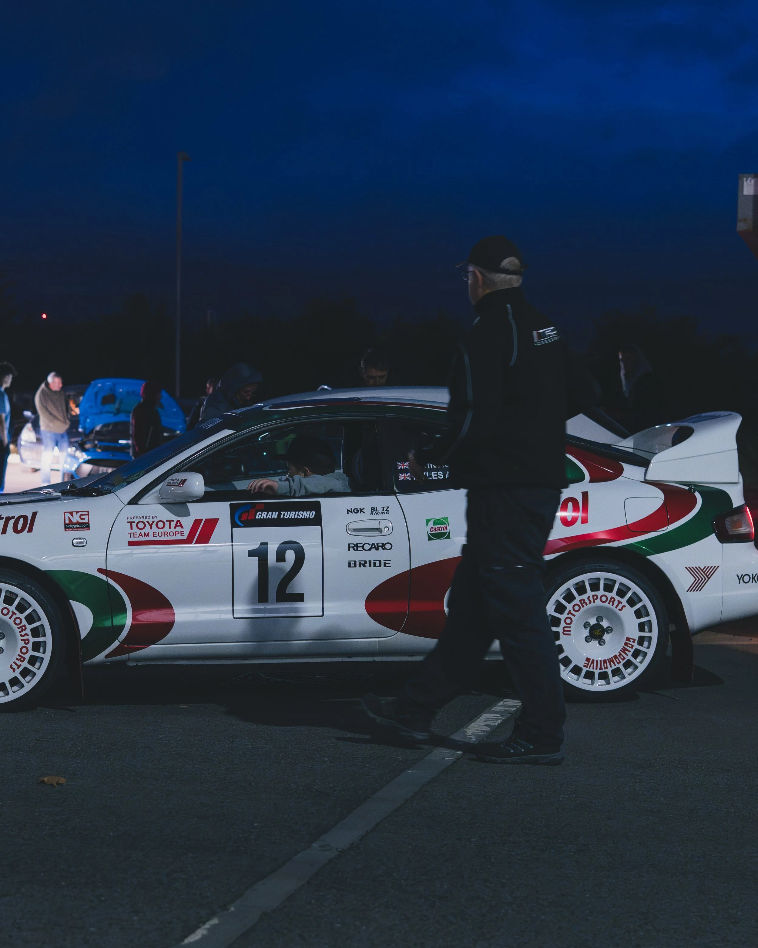 A race car with the number 12 and branded with Toyota Team Europe, NGK, Castrol, and other logos, parked at a racing event at night. A person is walking past the car, and other people are seen in the background near open car hoods.