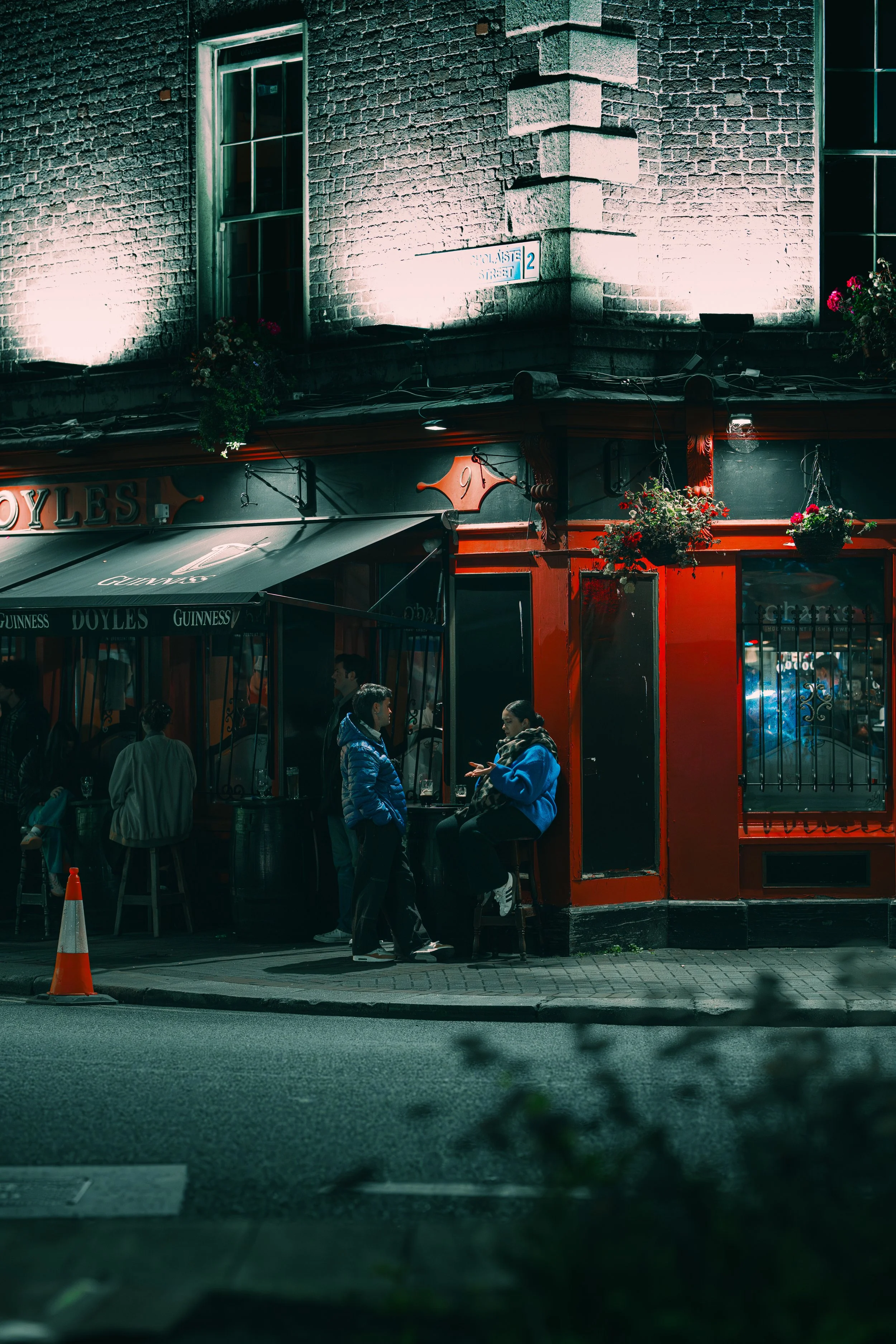 Nighttime scene outside a pub with people conversing at the sidewalk. The pub has a red exterior with hanging flower baskets and a Guinness sign. A cone is on the street, and a brick building with lit windows is in the background.