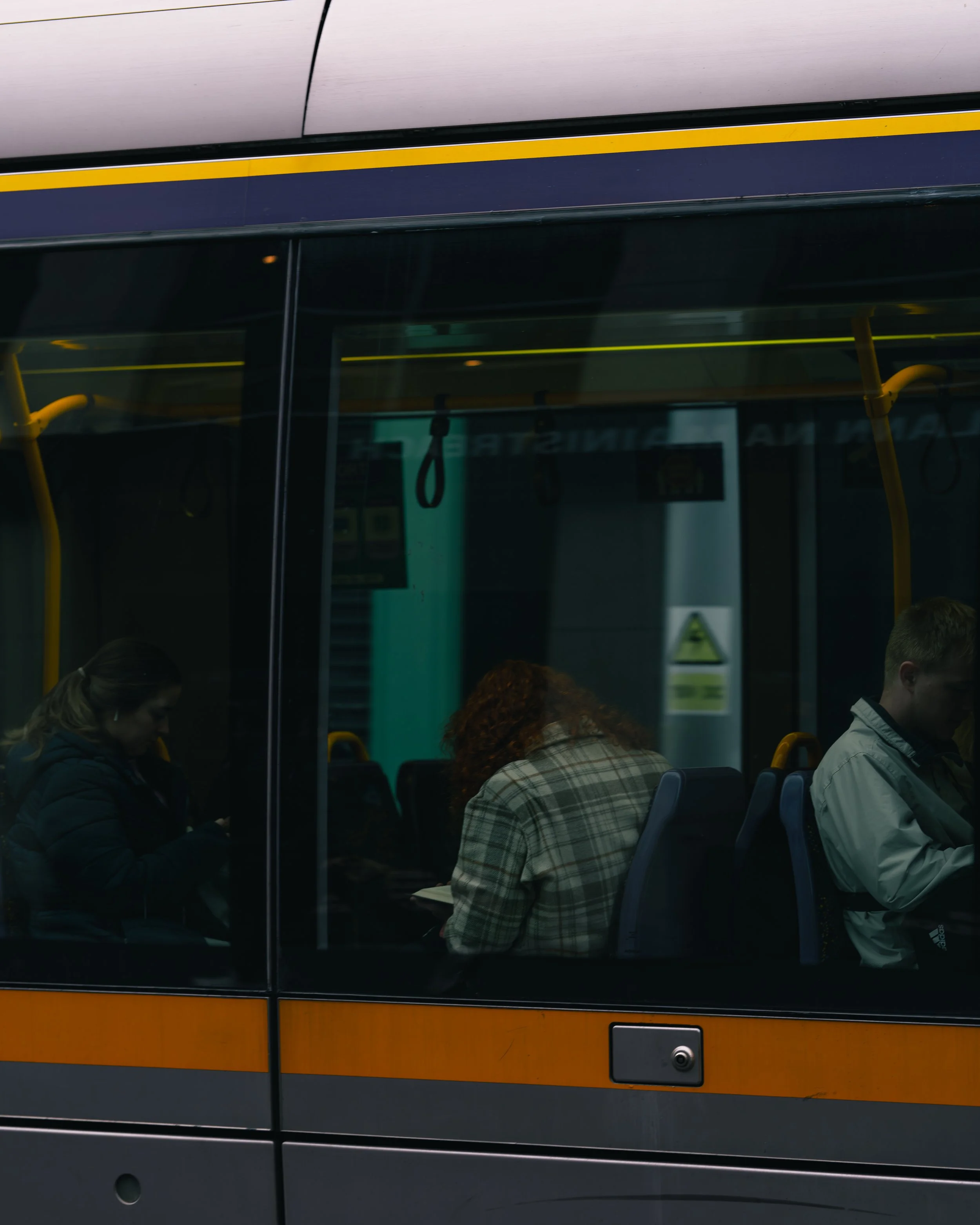 Interior view of a bus with three seated passengers, two women and one man, visible through the window, all engaging with their devices.