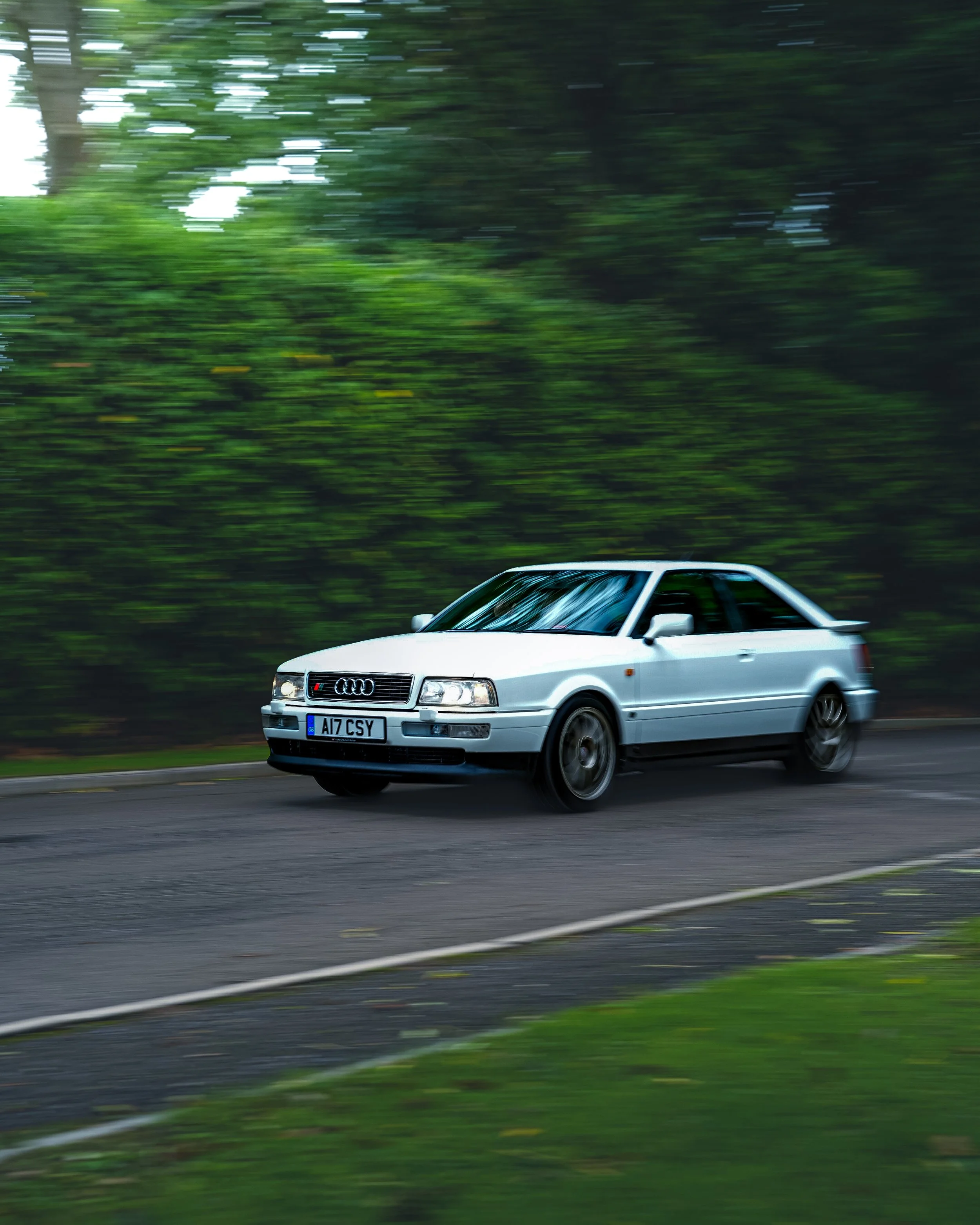 A classic white Audi coupe driving on a road surrounded by green trees, with motion blur indicating high speed.