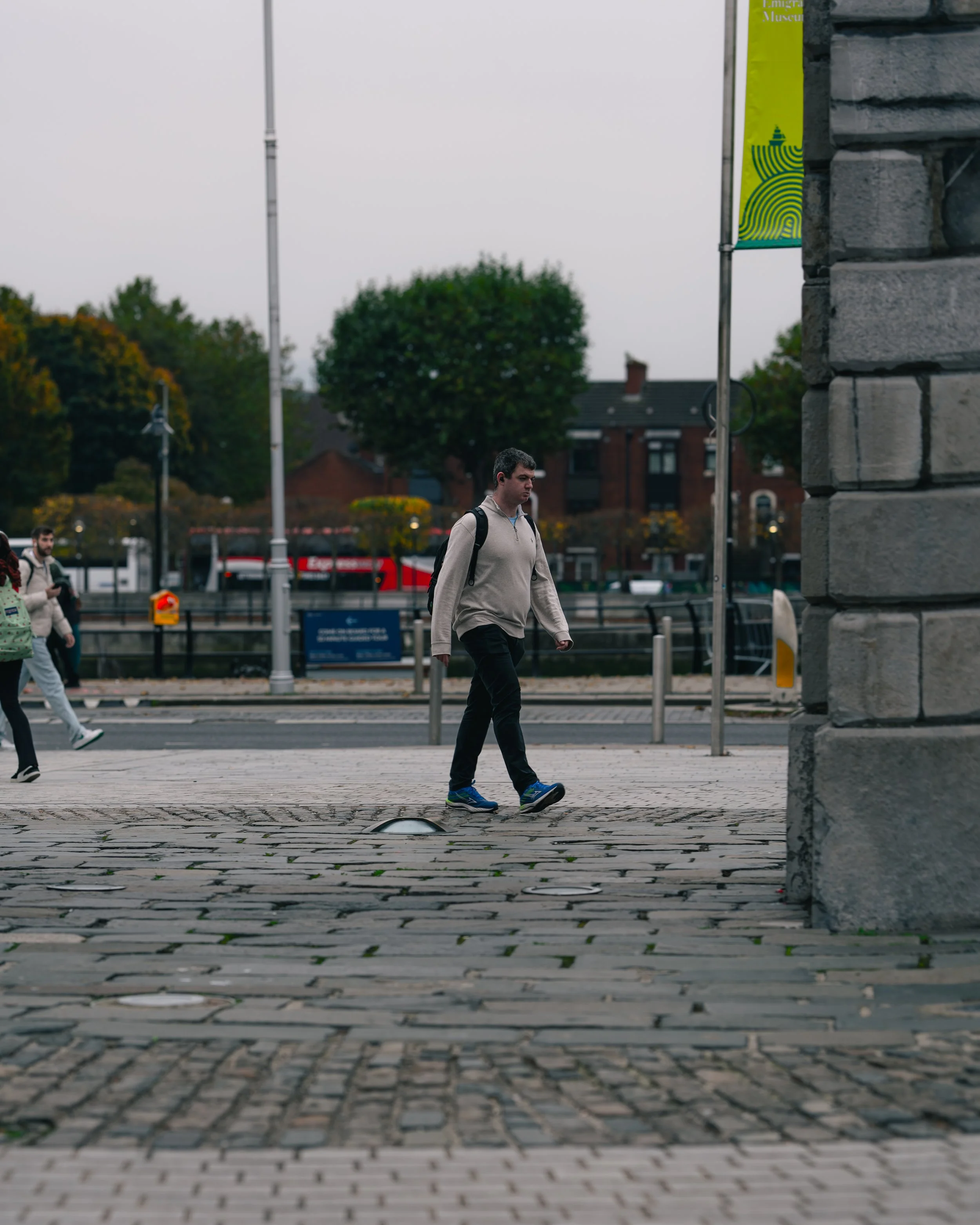 A man walking across a cobblestone street with a backpack, with other pedestrians nearby and trees in the background.