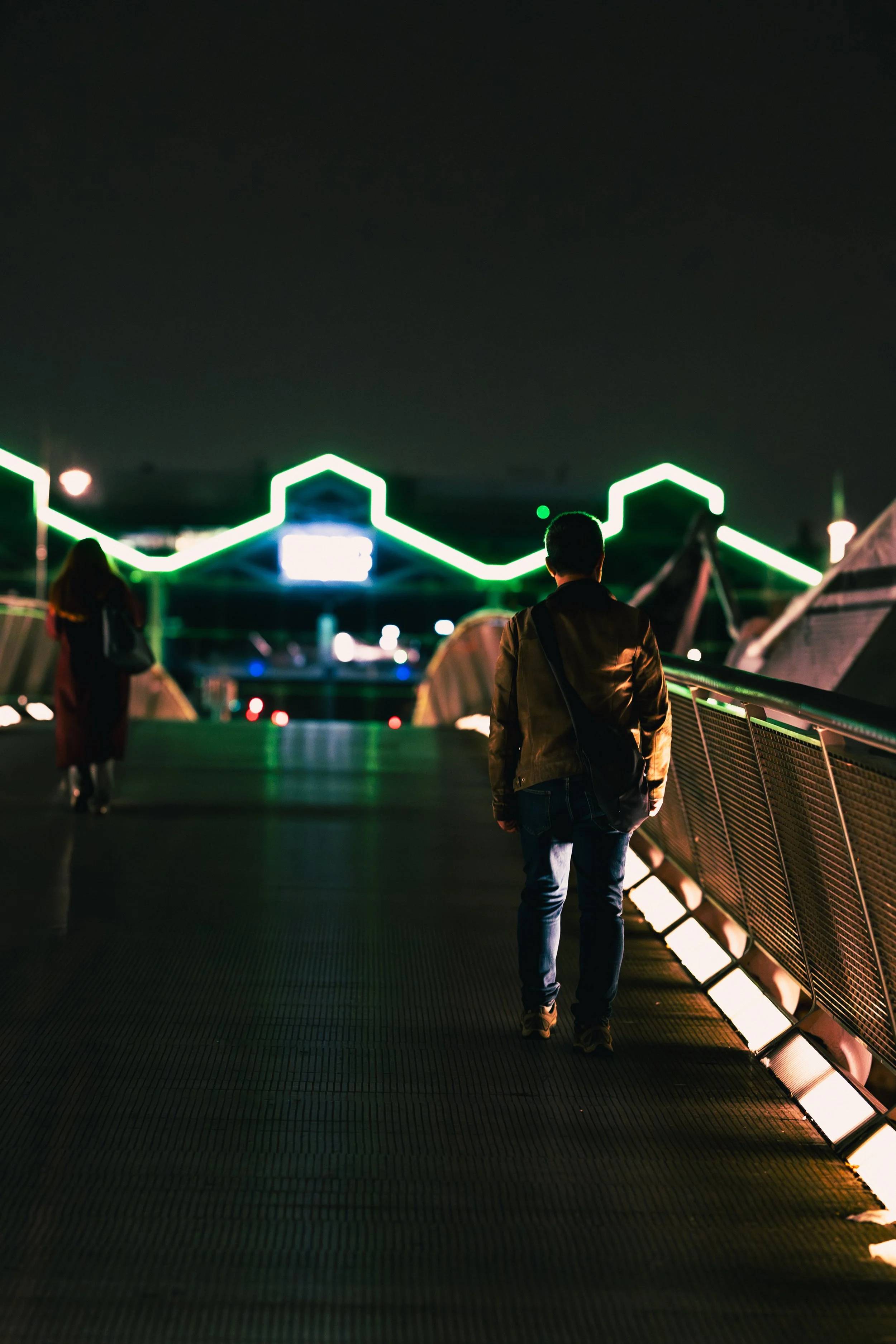 A man walking on a bridge at night with neon green lights and tents on the side, with a woman walking ahead in the distance.
