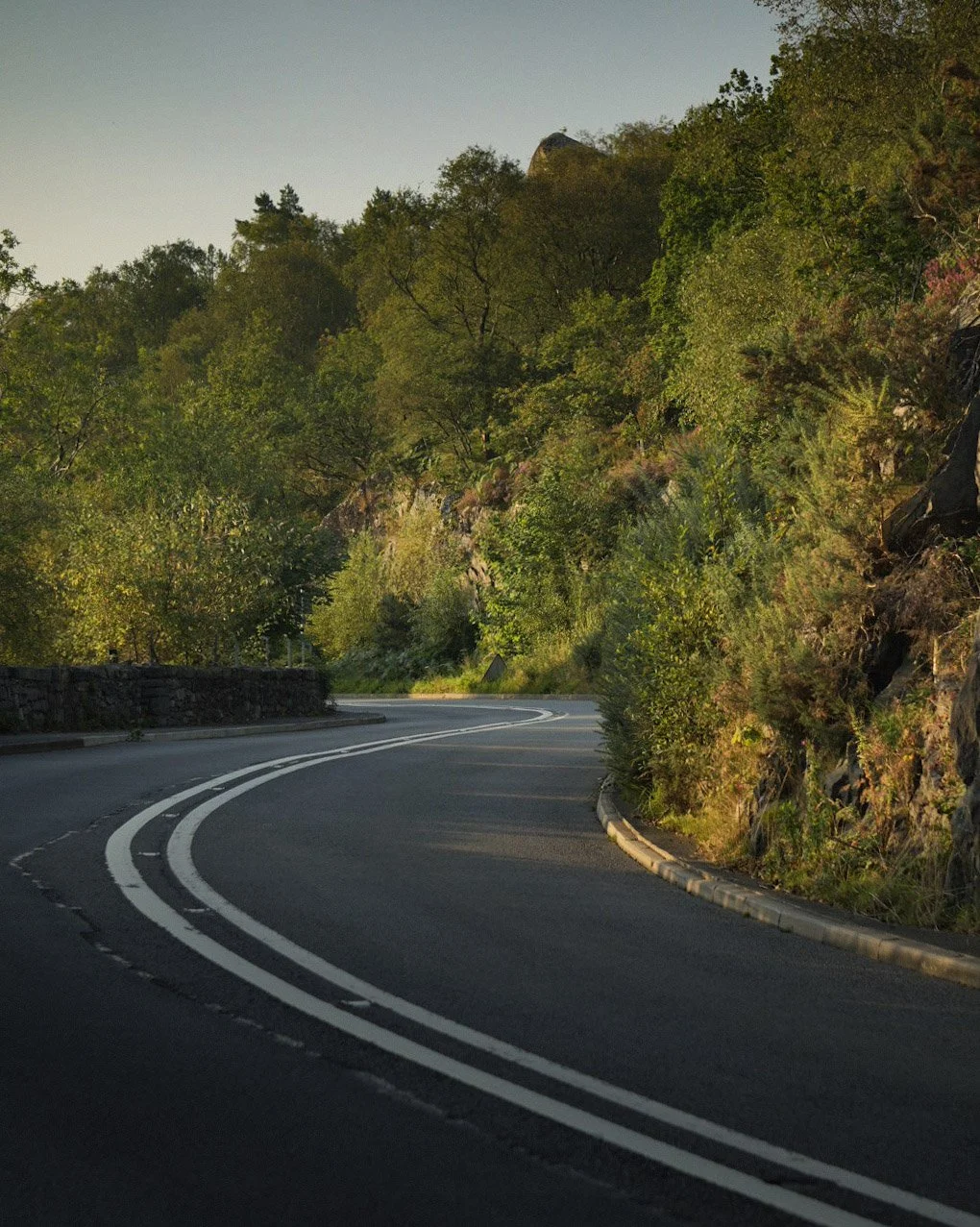 A winding two-lane mountain road bordered by trees and rocky cliffs, with a stone guardrail on one side, under a clear sky.
