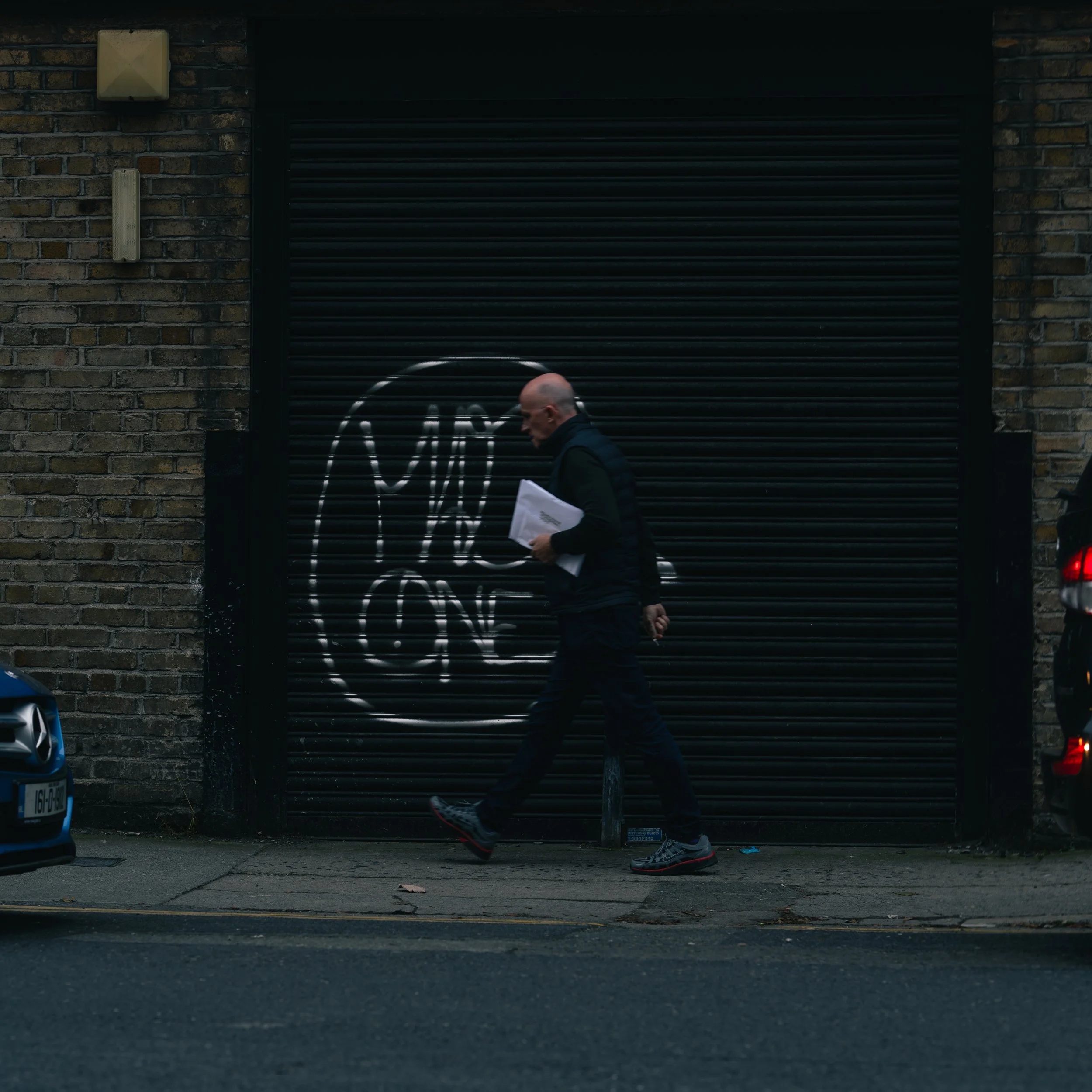 A man walking on a city sidewalk in front of a black garage door with white graffiti. He is holding papers and wearing dark clothing and sneakers.