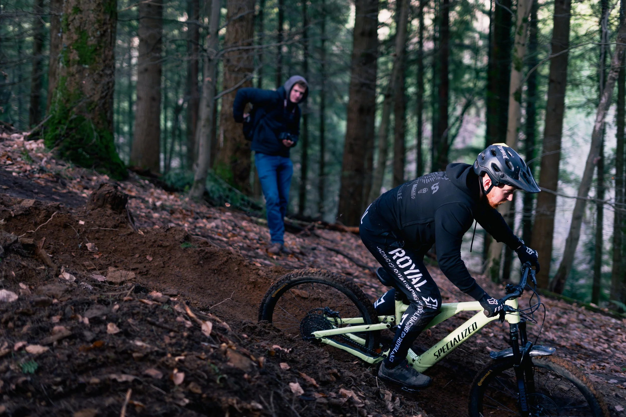 A person riding a mountain bike downhill on a forest trail, wearing a helmet and black gear, while another person in a hoodie watches from the background.