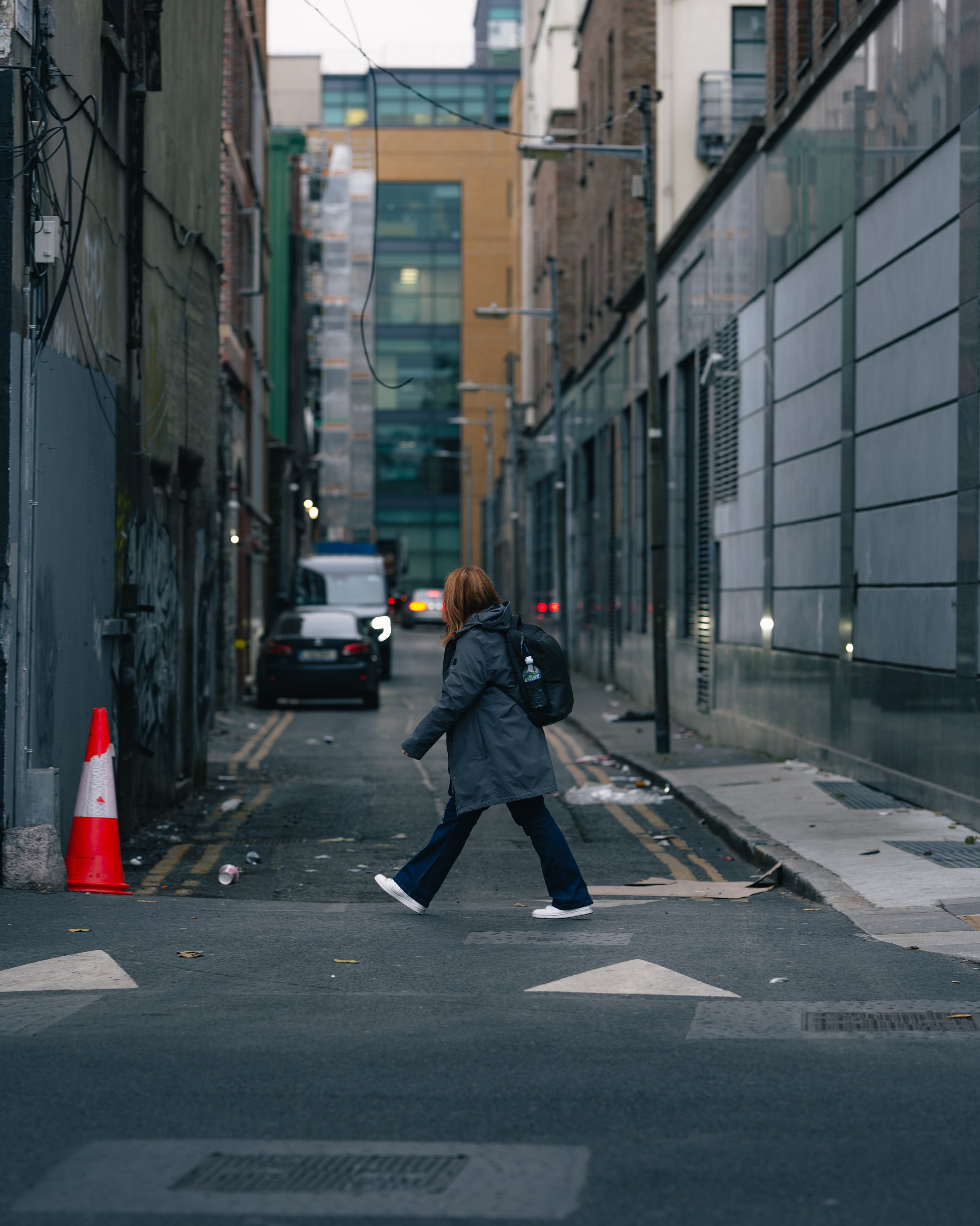 Person crossing the street in an urban alleyway with tall buildings on both sides, parked cars, and a traffic cone on the sidewalk.